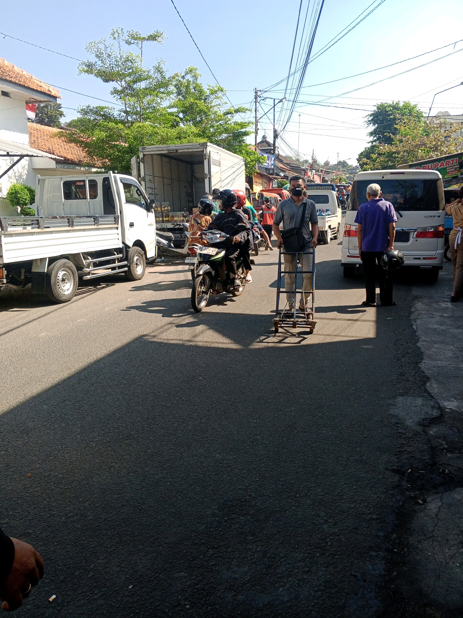The image depicts a street scene in Wonogiri, Indonesia, under a clear sky. In the foreground, a man wearing a gray shirt and black pants walks with a wheeled cart. To his left, a white truck is parked, its bed containing several large bottles of what appears to be cooking oil. Behind the truck, another white truck with an open back is visible. Further down the street, a white minivan and other vehicles are present. Several individuals, some on motorcycles and others on foot, are scattered throughout the scene. The environment is characterized by asphalt roads, sidewalks, buildings, and overhead electrical wires, suggesting an urban or semi-urban setting. The overall impression is a snapshot of daily life and commerce on a street in Indonesia.