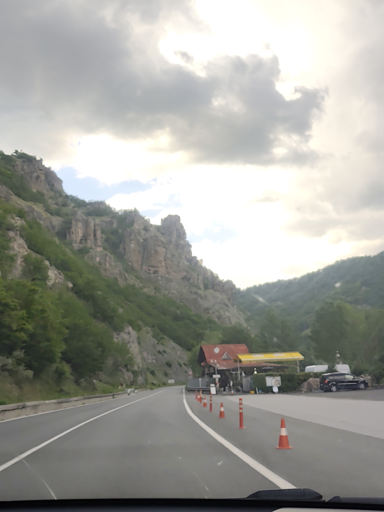 The image shows a highway passing through a mountainous region near Bukovo, Bulgaria. The highway has a white line separating lanes. On the right side of the road, there is a gas station or roadside stop with a red-roofed building and yellow awning. Several orange traffic cones are positioned along the white line, diverting traffic. A dark-colored minivan is parked near the building. Mountains covered in vegetation are visible on both sides of the road, with a notable rocky cliff on the left. The sky is overcast. The image is taken from inside a vehicle, with the dashboard visible at the bottom of the frame.