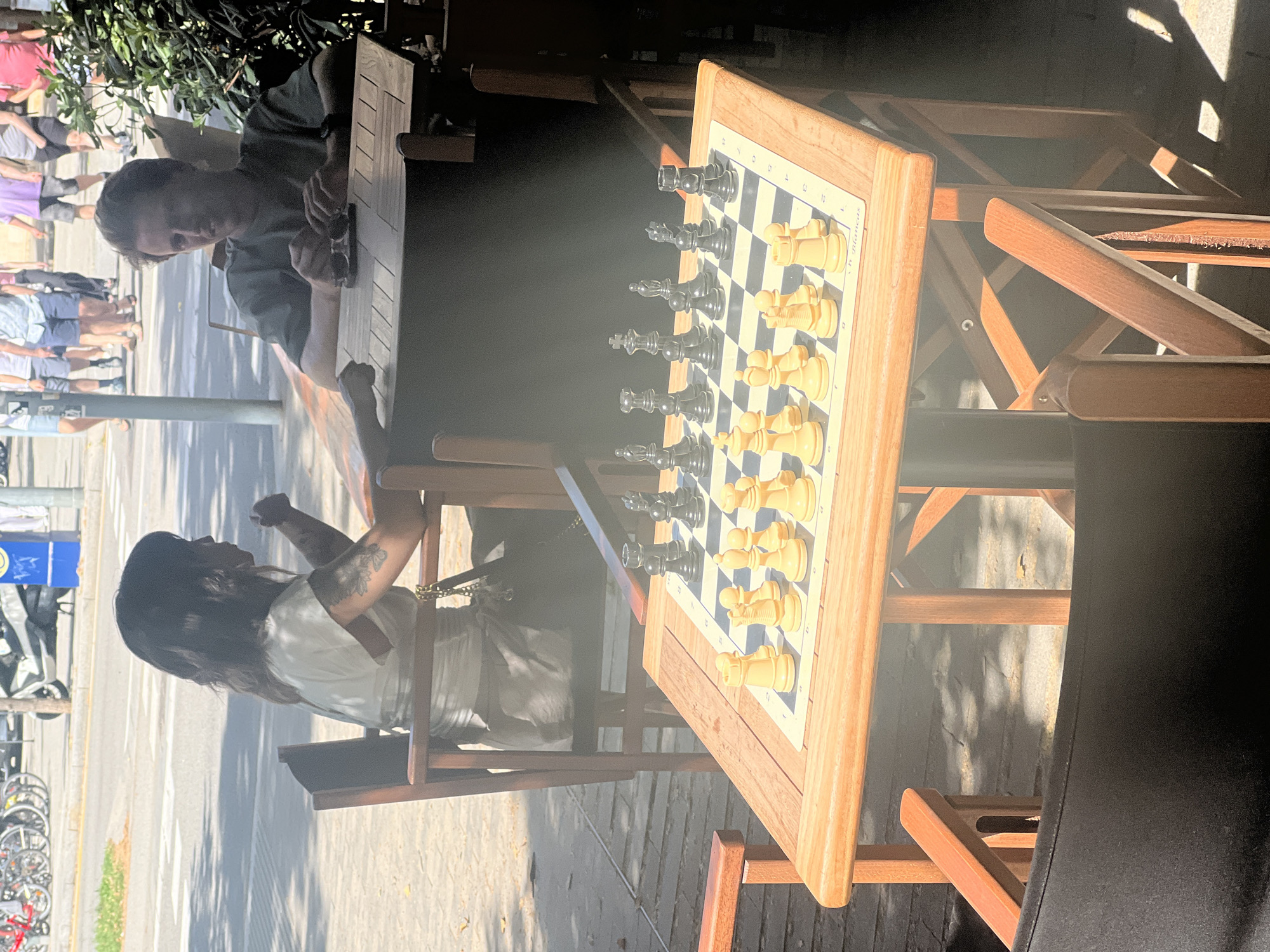 In Barcelona, Spain, at an outdoor cafe on a sunny day, a chess game is underway between a woman and a man. The woman, with dark hair and tattoos on her arm, is seated and looking away from the camera. The man, with light brown hair, is seated opposite her, looking towards the camera and holding sunglasses. A wooden chess table is positioned between them, displaying a game in progress with both white and black pieces set up. The cafe is situated on a tiled patio, with wooden chairs and tables visible. In the background, people are walking and a street with bicycles and a scooter can be seen. The scene suggests a leisurely afternoon activity in a bustling urban environment.