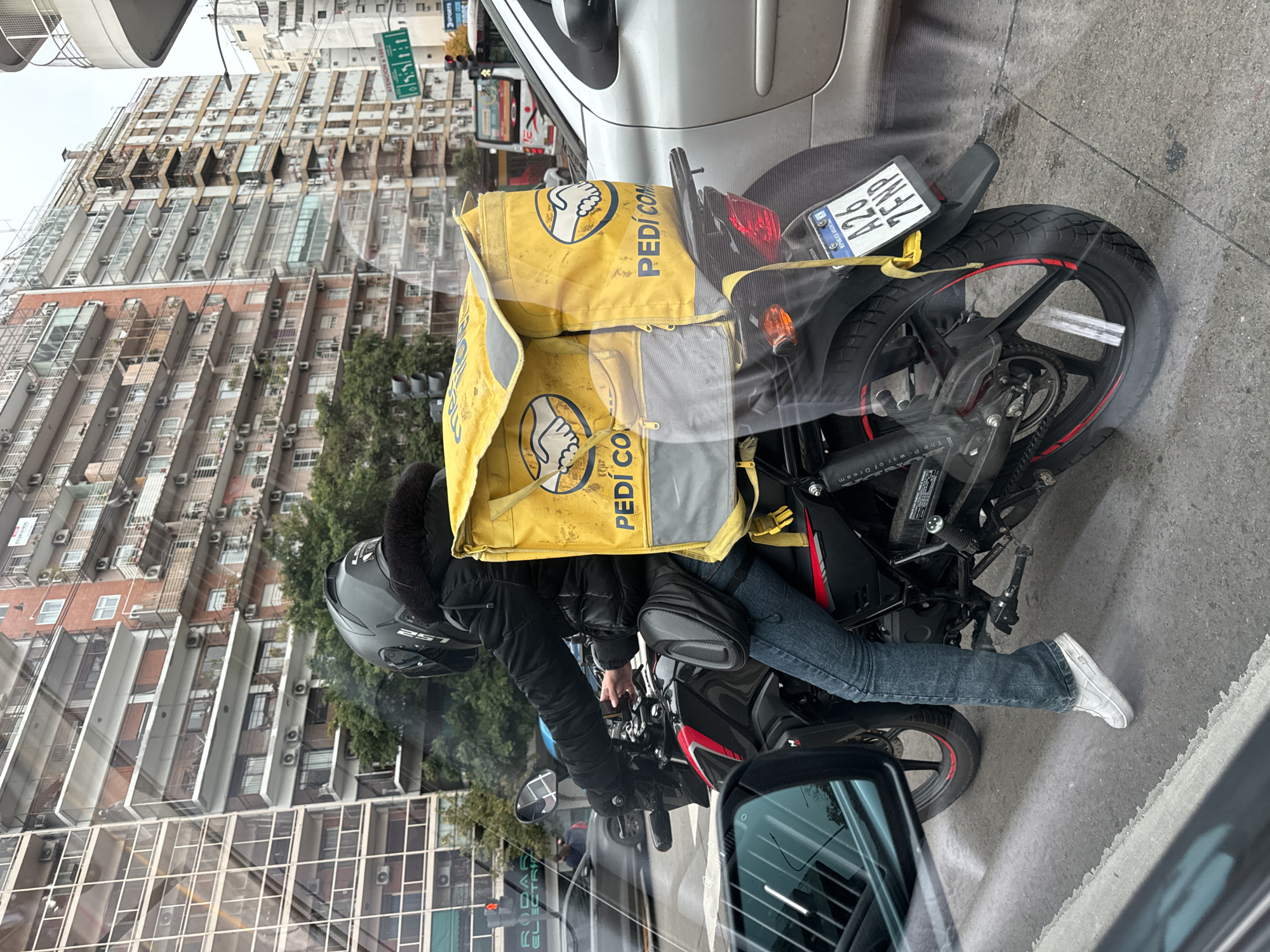A pedestrian view, through a vehicle window, shows a motorcyclist with a yellow "Pedidos Ya" delivery bag on the bike, stopped on a street. The motorcycle is black with red accents, and the license plate reads "A26 7FNP". The rider wears a gray helmet, black jacket, blue jeans, and white sneakers. The background includes apartment buildings, trees, and traffic signals, all consistent with an urban environment, specifically, the city of Buenos Aires, Argentina.