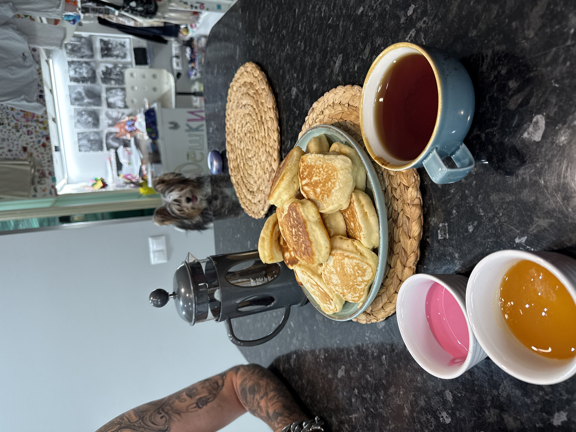 The image depicts a breakfast scene set on a dark granite countertop, likely in a kitchen. In the foreground, two small white ramekins hold pink and orange liquids, possibly jams or syrups. To the right, a blue ceramic mug filled with dark tea sits. Behind these, a plate of golden-brown pancakes is presented on a woven placemat. A French press coffee maker, dark gray with a glass carafe, is positioned to the left of the pancakes. Further back, a small dog with brown and white fur sits attentively. The background features a desk area with artwork and decorative items, suggesting a home environment. The overall impression is of a casual, domestic setting. The requested location of Sochi, Russia, is not directly inferable from the visual evidence.