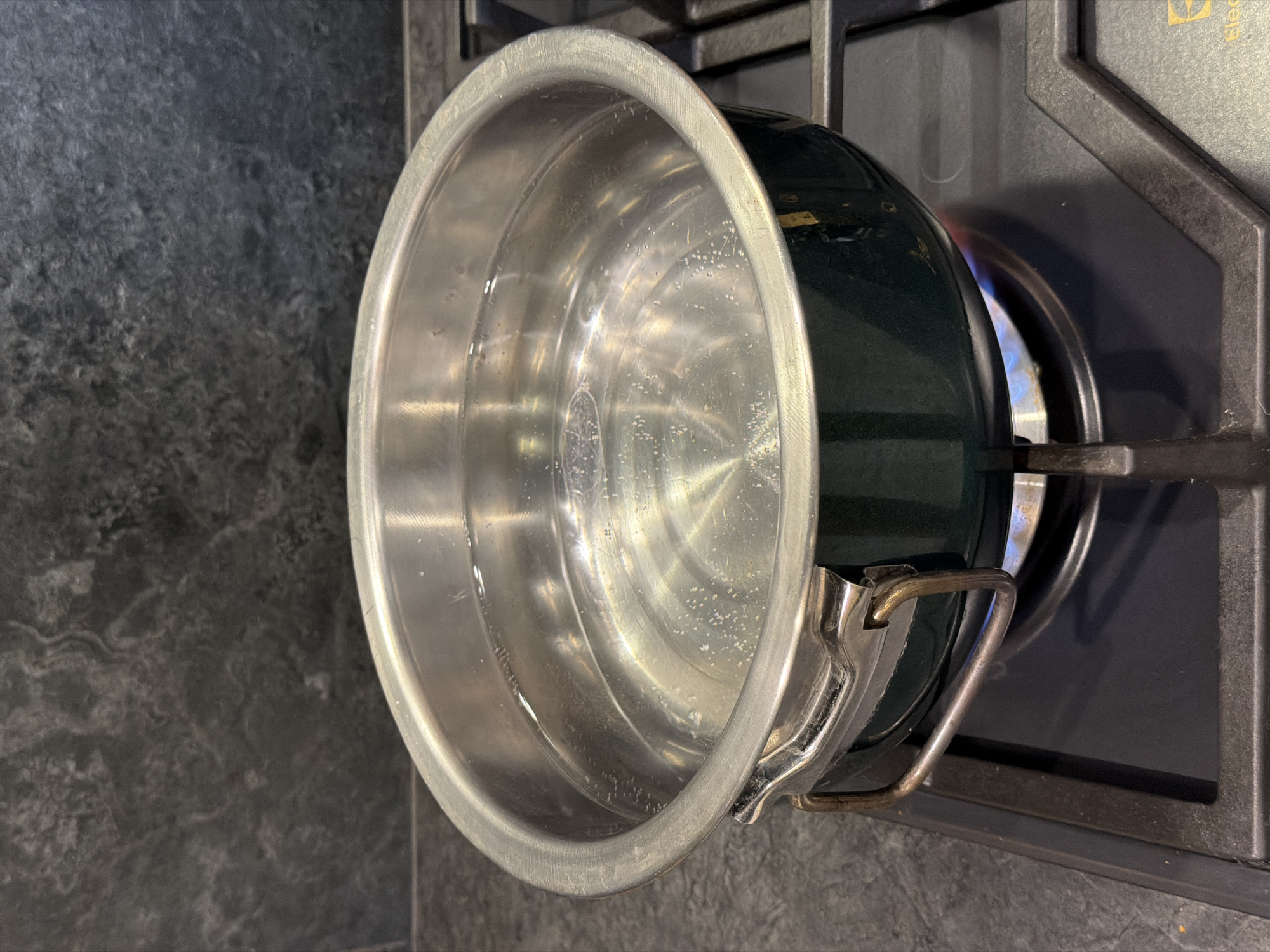 A pot of water is being heated on a gas stove. The pot is made of stainless steel with a dark green exterior. The water is clear and has small bubbles forming on the bottom and sides, indicating it is starting to boil. The stove top is black with a dark gray speckled backsplash behind it. The flame from the burner is visible underneath the pot, emitting a blue light. There are no people present in the image.