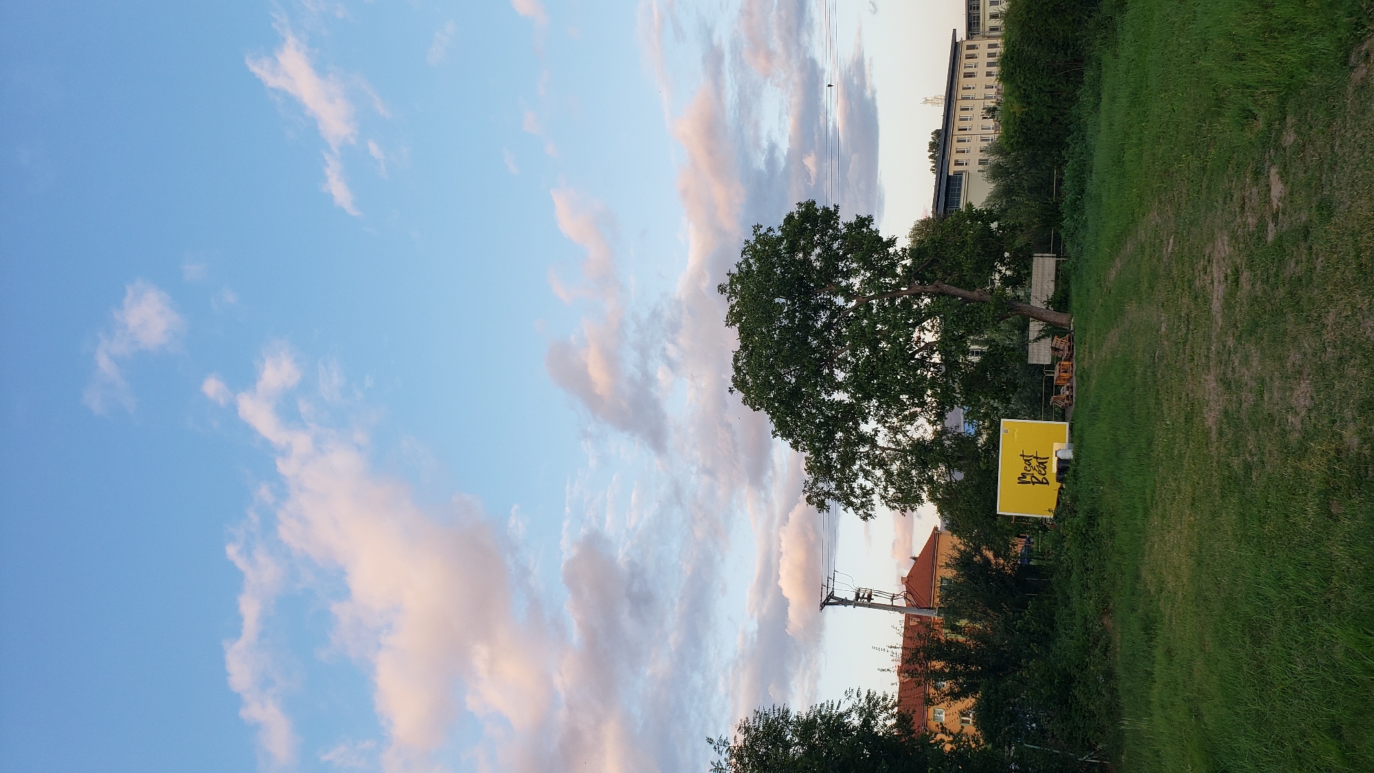 This image depicts an outdoor scene in Prešov, Slovakia, likely at dusk. The sky is a soft blue with scattered pink and white clouds. In the foreground, a grassy area with a dirt path leading towards the center is visible. To the left, a large tree with dark green foliage dominates the mid-ground. Behind the tree and to the left, there's a yellow sign with "Meat Beat" written in black script. Further back, a residential building with a red roof and several trees can be seen. To the right of the yellow sign, there are some wooden chairs and tables. A long, multi-story building with a light-colored facade is visible in the background on the right. Power lines and poles are also present in the scene. The overall atmosphere is calm and serene.