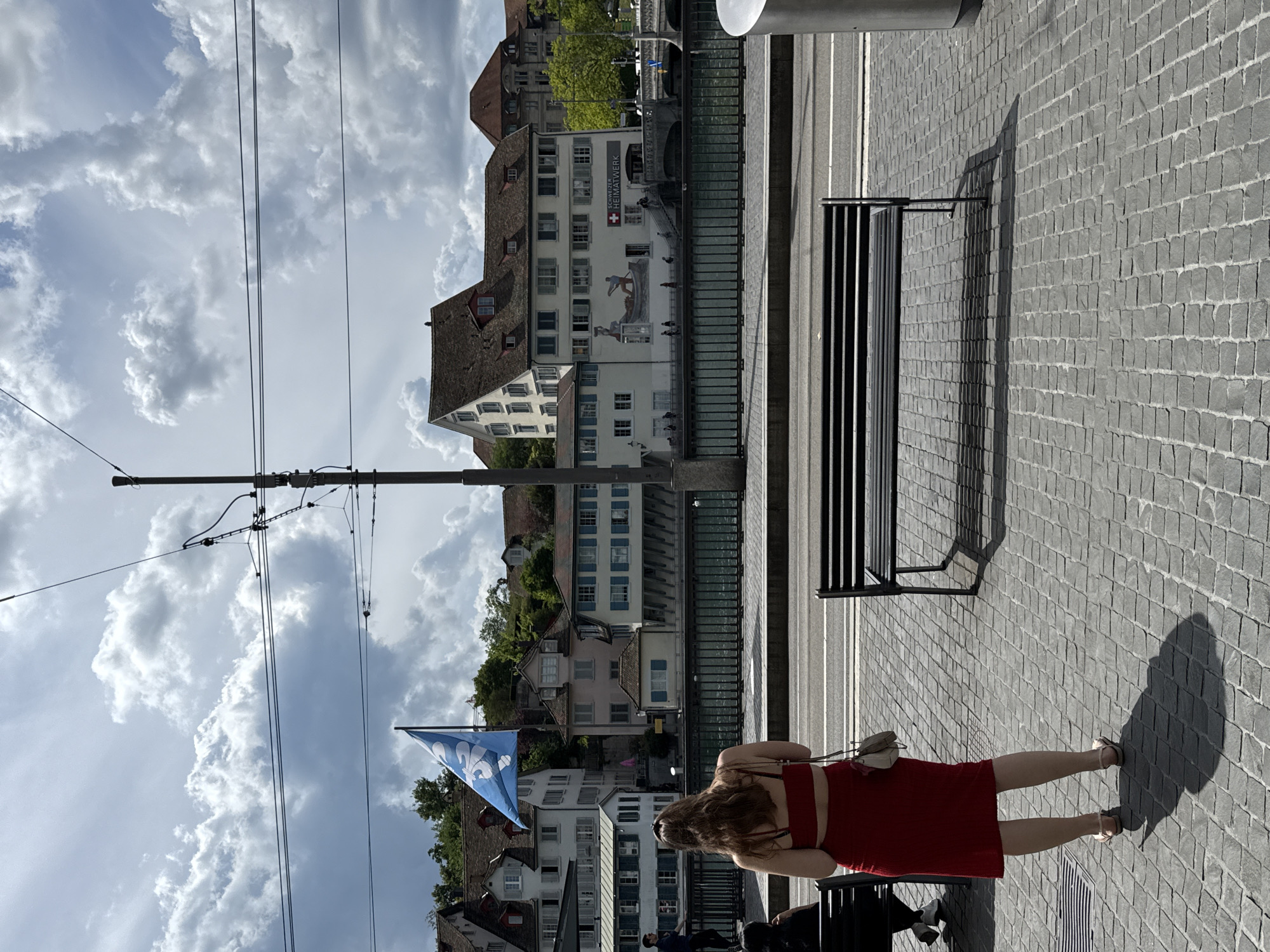 The image captures a daytime view of a waterfront promenade in Zurich, Switzerland. The foreground features a woman from behind, wearing a red dress and sandals, standing on a cobblestone pavement. Next to her, to the left, is a black park bench with a shadow cast on the pavement. Further to the left, another park bench is partially visible, occupied by a person. In the middle ground, a river or canal is visible, bordered by a railing. Across the water, a row of buildings lines the embankment, characterized by traditional architecture with pitched roofs and multiple windows. A prominent blue flag with a white emblem flies from a pole in front of the buildings. In the background, more buildings and trees are visible, suggesting an urban setting with greenery. The sky is partly cloudy, with dramatic cloud formations. Power lines and a utility pole are present in the upper portion of the image. One of the buildings across the river has text that reads "SCHWEIZER HEIMATWERK" and a Swiss flag emblem.
