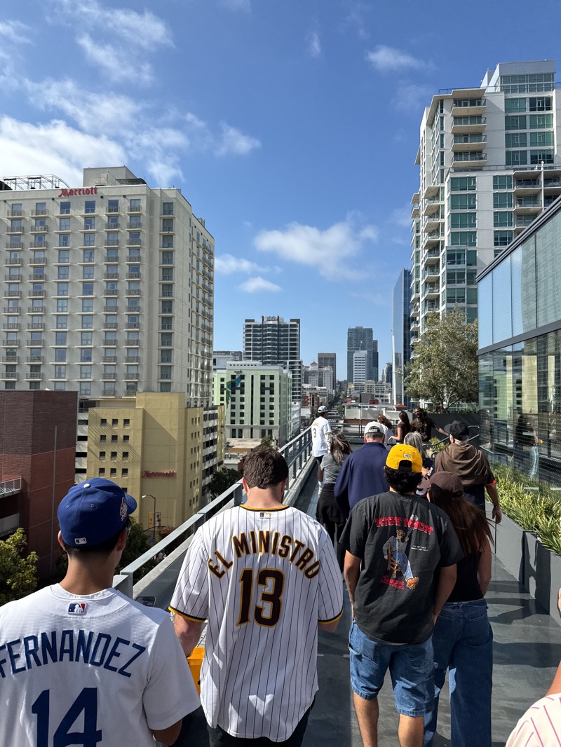 A medium, eye-level, outdoor shot shows a group of people walking on an elevated walkway with a view of the city skyline of San Diego, United States. The sky is blue with scattered clouds. The foreground is dominated by the backs of several people, most wearing sports jerseys and casual clothing. The person in the front on the left wears a white jersey with “FERNANDEZ” and the number “14”. Next to him, another person wears a white baseball jersey with pinstripes, “EL MINISTRO” written on the back, and the number “13”. Another person in the middle wears a black t-shirt with a yellow cap.
Behind the group, the buildings of downtown San Diego form a backdrop. On the left, a tall, light-colored building is identified by the “Marriott” sign. Other skyscrapers and buildings of varying heights are visible in the background. The walkway has a metal railing and greenery on the right side.
