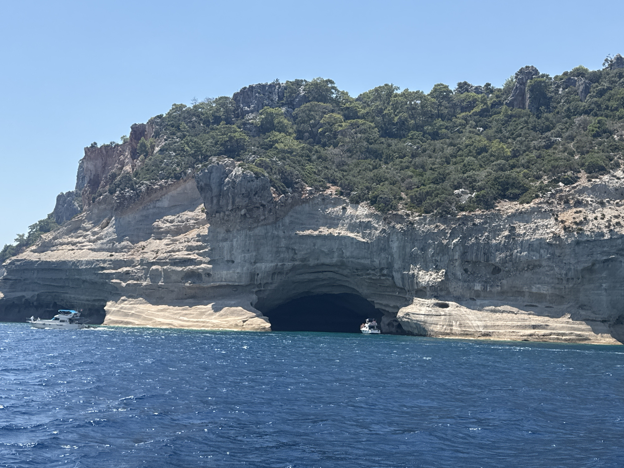 An aerial view shows a wide, dark cave entrance in a towering, grey cliff face along the Mediterranean Sea, near Kemer, Turkey. The cliff is covered in lush green trees and vegetation near the top, with stratified rock formations visible on its lower half. Two white boats are present: one smaller, white vessel with a blue canopy is moored near the left side of the cave, and a larger, white tour boat with a blue stripe is entering the cave's opening on the right. The sea is a deep, rich blue with gentle waves. The sky is clear and bright blue.