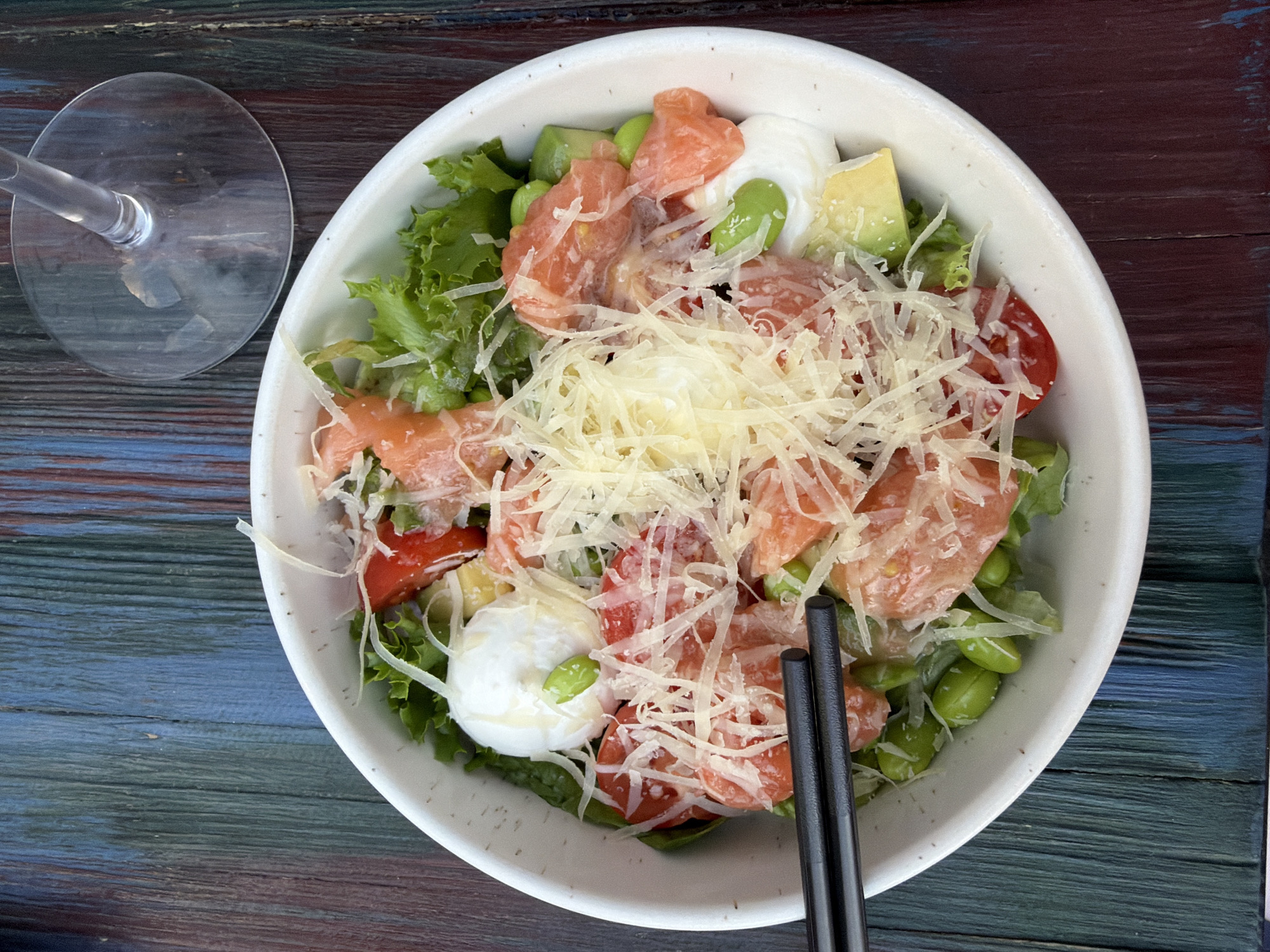 A close-up, overhead shot captures a colorful salad served in a white bowl, resting on a rustic, dark wooden table. The salad is a vibrant mix of ingredients including chunks of pink salmon, halved cherry tomatoes, bright green edamame beans, sliced avocado, and crisp lettuce leaves. A generous topping of shredded cheese crowns the dish. Two black chopsticks are positioned on the right side of the bowl, suggesting the salad is ready to be eaten. To the left of the bowl, the base of a clear wine glass is visible, adding to the dining scene. The wooden table has a weathered appearance with visible grain and variations in color, appearing to be painted in shades of dark blue, green, and red. The lighting suggests it is daytime, likely indoors or in a shaded outdoor area, as there are no harsh shadows. No people are visible in the frame, but the presence of the salad and glass implies a meal is about to be enjoyed. There is no visible text in the image. The setting appears to be a casual dining environment, potentially a restaurant or a home dining table. The overall impression is one of fresh, appetizing food presented in a rustic setting.