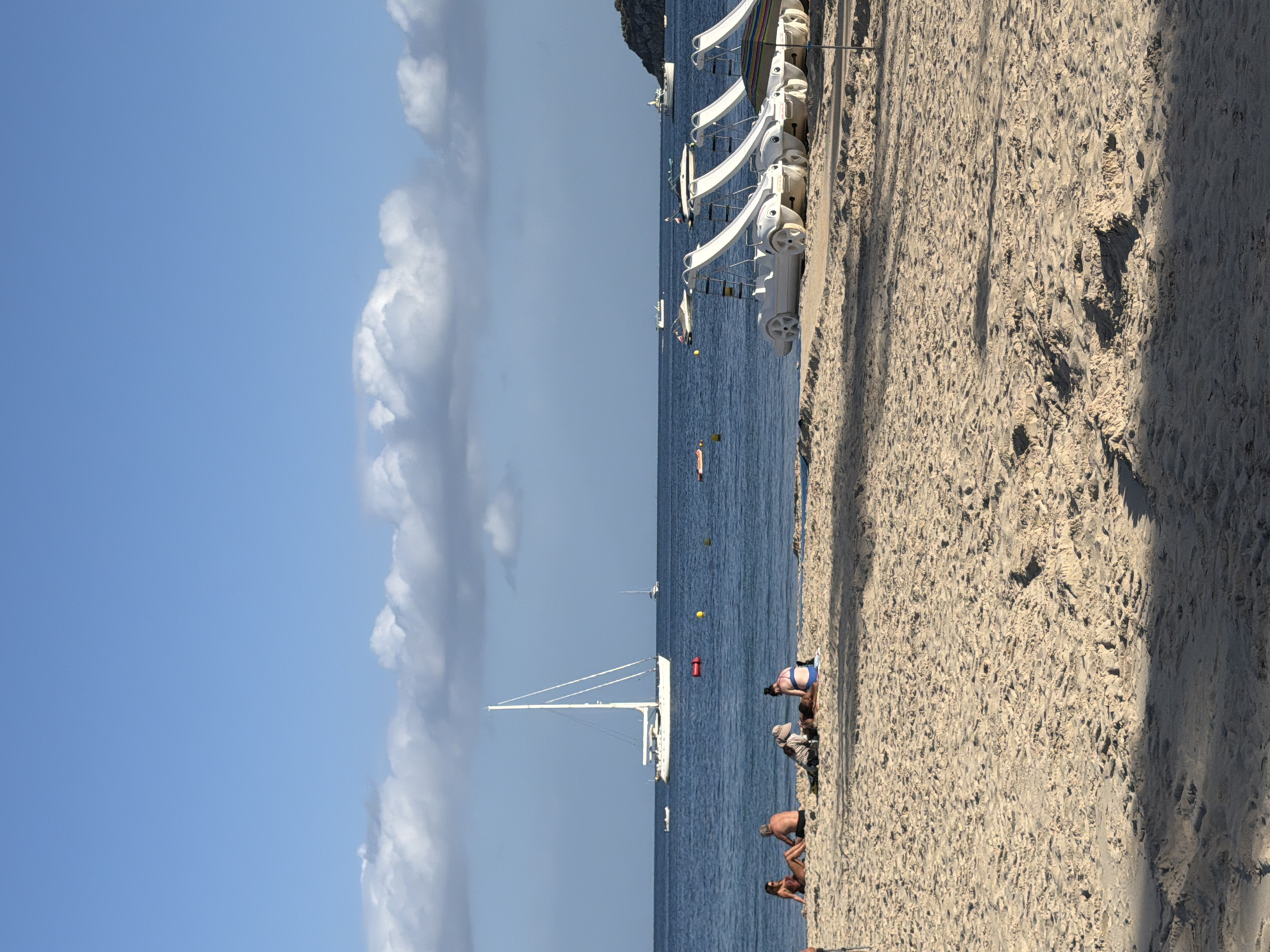 In Palmanova, Spain, a sandy beach is depicted under a bright blue sky with scattered white clouds. Several people are relaxing on the beach in swimwear. Some are seated, while others are lying down. In the shallow water near the shore, a line of white pedal boats with slides are parked. The sea stretches out to the horizon, where a large white sailboat is anchored, accompanied by a few smaller boats and a distant landmass. The sand is textured with footprints and shadows from unseen objects.