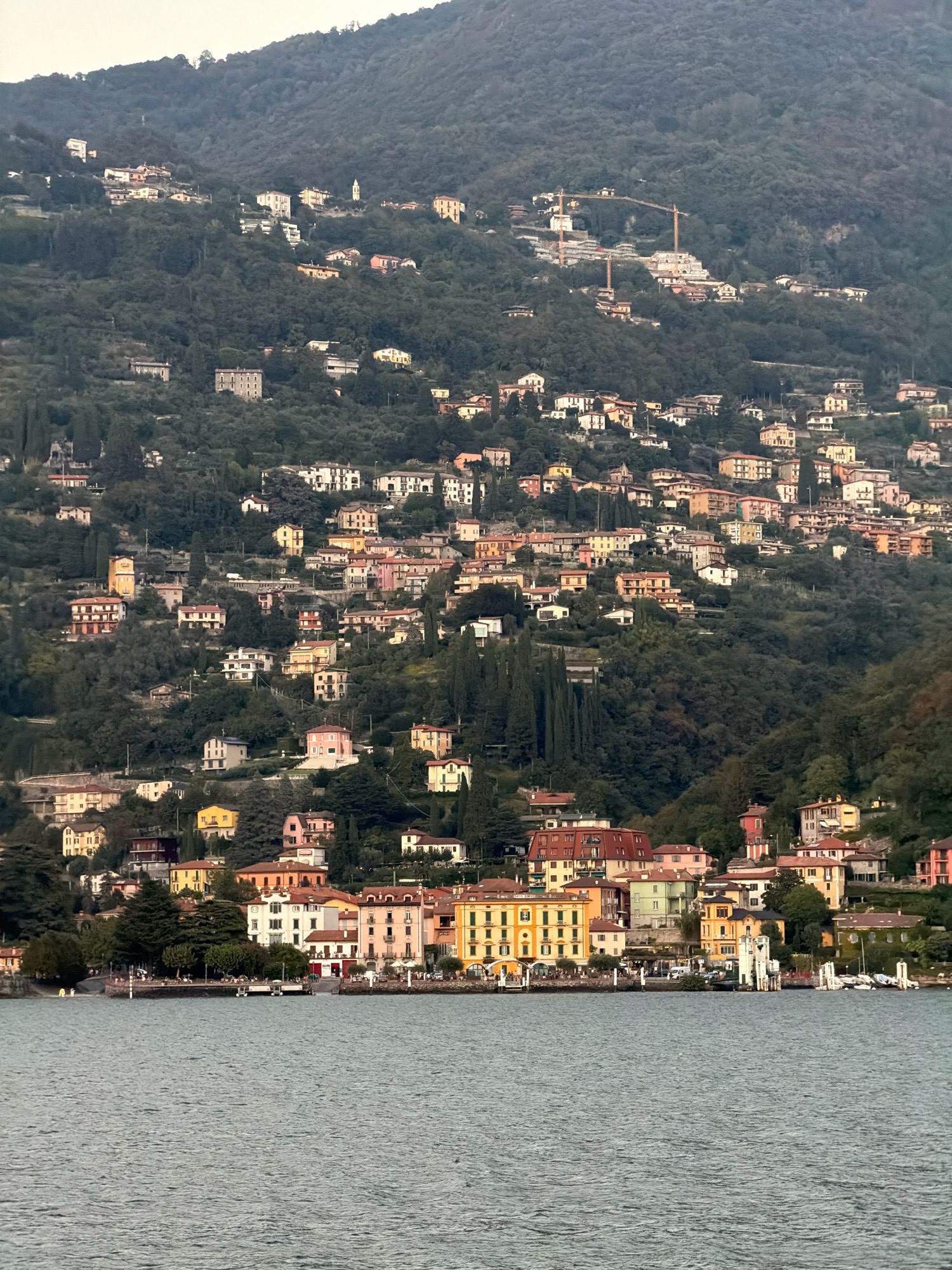 The image depicts a waterfront view of the town of Varenna, Italy, situated on the slopes of a densely forested hill. The foreground is dominated by a body of water, likely Lake Como, with a slightly rippled surface reflecting the overcast sky. Along the shoreline, a vibrant collection of buildings with colorful facades, varying in architectural style and height, are clustered together. These buildings, painted in shades of yellow, pink, orange, green, and white, feature multiple stories and prominent windows, suggesting residential and commercial use.
Several boats are moored at a dock to the right of the frame, with a white, tower-like structure standing near the water's edge. A promenade or street runs along the waterfront, lined with trees, and appears to be populated by a few individuals, though they are indistinct due to the distance. Some outdoor seating areas are visible.
The mid-ground and background are characterized by the steep hillside, densely covered in lush green foliage. Scattered amongst the trees are more houses and buildings, climbing higher up the slope. Some of these appear to be older structures, while others suggest ongoing construction, evidenced by visible cranes in the upper right section of the hillside. The overall impression is of a charming, densely built Italian lakeside town nestled into a dramatic natural landscape.
