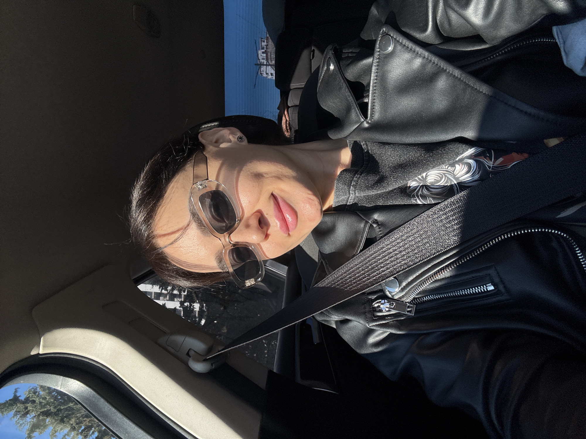 The image is a selfie taken inside a car. A woman wearing sunglasses and a black leather jacket is wearing a seatbelt. Outside the car window, there is a view of some buildings and greenery. While the exact building is difficult to identify, given the context it is likely a building in Tbilisi, Georgia.