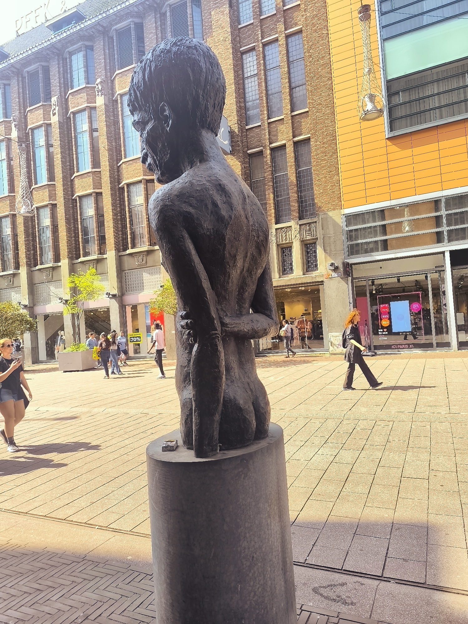 The image shows a bronze bust of a man's torso from behind, positioned on a cylindrical pedestal in an outdoor pedestrian area. The bust is dark and textured, depicting an emaciated figure with its arms crossed behind its back. The background features a multi-story brick building with numerous windows and some modern orange-tiled architecture with large glass windows. Several people are visible walking in the pedestrian area, some are in groups, while others are alone. One woman with red hair is walking across the foreground from right to left. Another woman in a black top and shorts is walking from left to right in the mid-ground. The ground is paved with light-colored rectangular tiles. This scene is located in the city of The Hague, Netherlands, likely on a shopping street or public square. The overall lighting suggests a bright, sunny day.