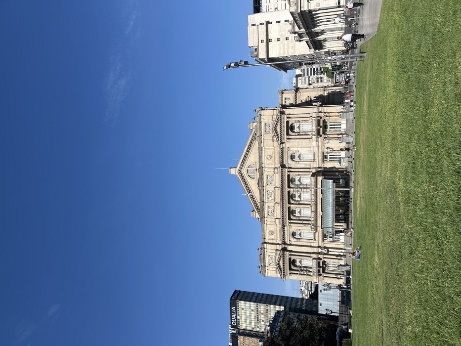 The Teatro Colón in Buenos Aires stands grandly beside a green lawn. Its ornate facade showcases intricate architectural details. Clear blue skies fill much of the frame. Adjacent buildings and a distant skyline complement the opera house. People are seen on the lawn and sidewalks, adding a sense of scale. Modern elements like a street light and a skyscraper blend with the classical architecture.