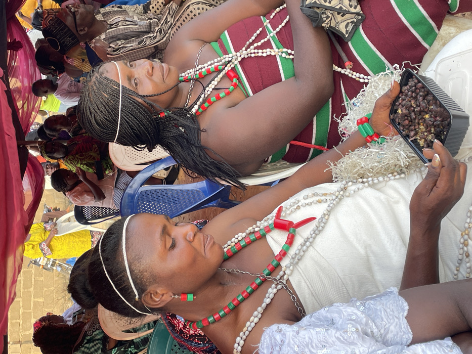 The image depicts an outdoor gathering or ceremony, filled with people, prominent among them two women adorned with traditional beads and attire. The overall lighting suggests bright daylight.
In the foreground, slightly to the right, a woman is seen wearing a white garment. Her head is adorned with a thin white headband, and her hair appears to be very short or shaved on the sides with some styled hair on top. She wears multiple strands of white and patterned beads around her neck and chest. A striking necklace with rectangular red and green beads, along with matching earrings, complements her attire. Her eyes are closed or cast downwards, giving her a serene or contemplative expression. She holds a black rectangular container filled with what appears to be dark-colored food, possibly beans or nuts, in her right hand. Her fingernails have a light polish or glitter. White, shredded decorative material, possibly tinsel or paper, is wrapped around her wrist and arm.
To the left of this woman, in the mid-ground, another woman is visible. She is dressed in a top or dress featuring maroon and green stripes. Her black hair is styled in long braids, pulled back and secured with a white band similar to the other woman's. She also wears multiple strands of white beads and a necklace of red and green beads, mirroring the style of her counterpart. Her gaze is directed upwards and slightly towards the viewer, with a calm and composed expression. Her arms are bent and resting in front of her.
In the background, numerous other individuals are visible, mostly seated on blue and white plastic chairs. They appear to be attendees or participants in the event, with some wearing patterned clothing. On the far left, a section of red, translucent fabric, possibly part of a canopy or tent, is visible, suggesting the gathering is held under shelter. The ground in some areas appears to be paved or tiled.
The prominent use of beads, vibrant colors (red, green, white, maroon), and distinct hairstyles suggests a cultural, traditional, or ceremonial event. The presence of food being held by one of the central figures could indicate a ritualistic offering, a shared meal, or a specific part of the celebration.