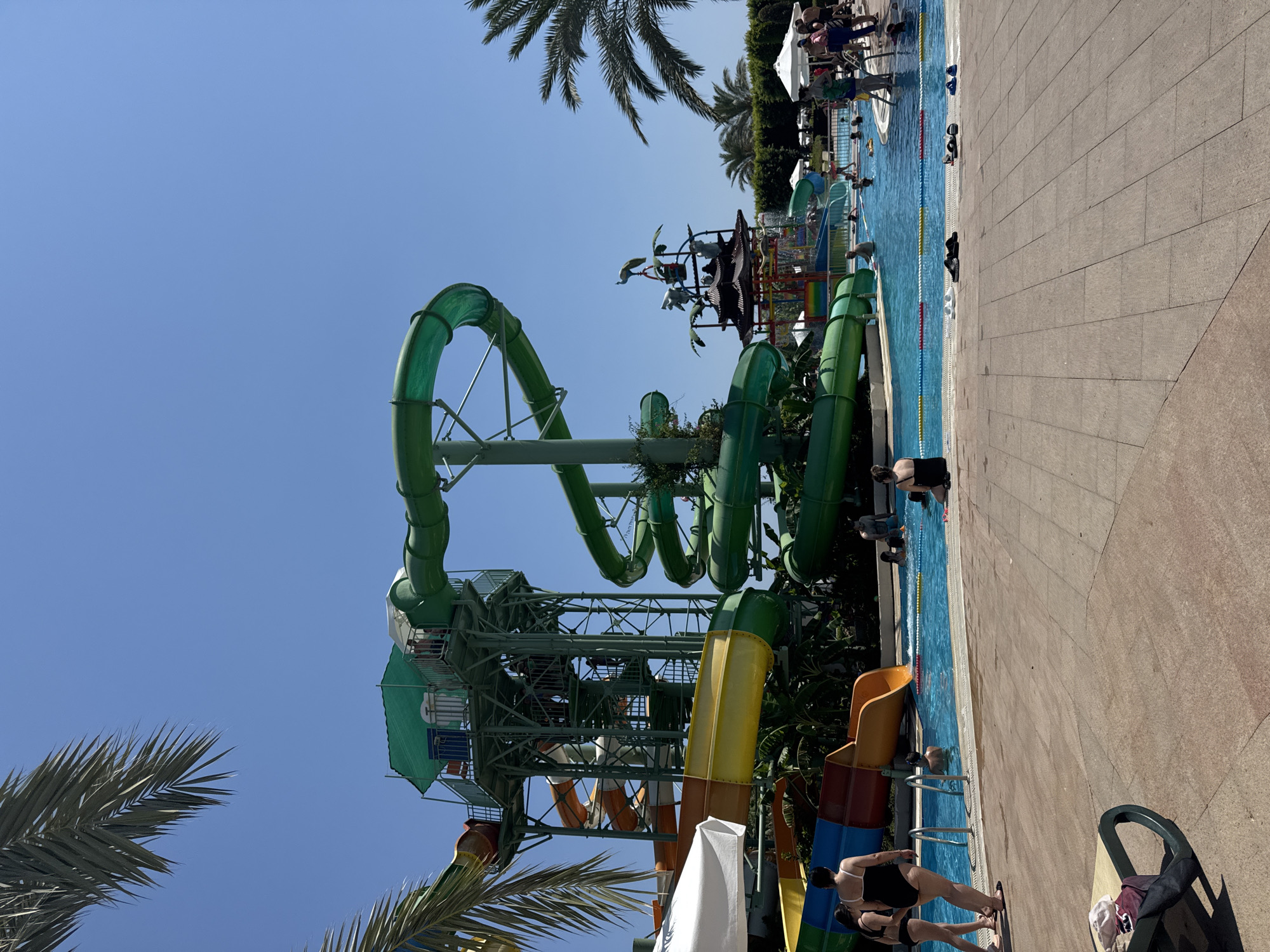 In Aksu, Turkey, a vibrant water park is captured on a sunny day. Dominating the scene are several large, colorful water slides. A towering green spiral slide curves downwards, juxtaposed with a yellow and orange slide descending from a multi-tiered structure. A smaller red and orange slide is also visible. The water park features a clear blue swimming pool where several people are seen relaxing and swimming. Along the pool's edge, lounge chairs and umbrellas are scattered. Palm trees frame the scene, adding to the tropical ambiance. People, dressed in swimwear, are scattered throughout the park, some entering the pool, others walking nearby. The environment is bright and cheerful, indicative of a recreational area.