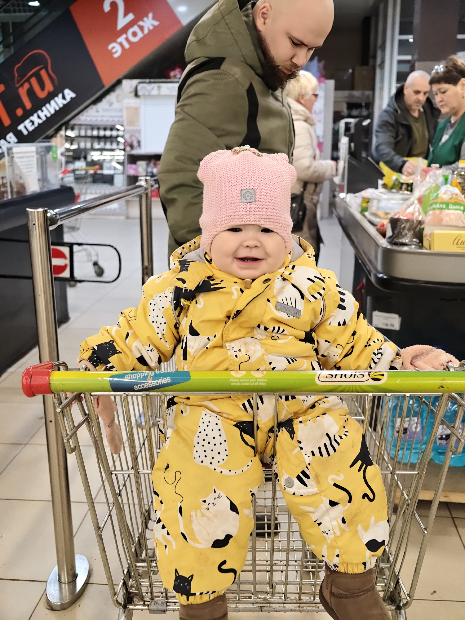 A young child wearing a yellow jumpsuit with a pattern of cats, a pink knit hat, and brown shoes, sits in a shopping cart. The child is smiling. A bald man with a beard, possibly the child's father, is standing behind the cart. An older woman with white hair is standing behind him. A cashier and a man are checking out items. The scene appears to be a grocery store in the city of Chaikovsky, Russia, as indicated by the signage in Russian in the background. A sign for electronics on the second floor is visible. The shopping cart has a green handle with red ends and a blue sticker on it. The floor is tiled. Bottled water is in the cart behind the child.