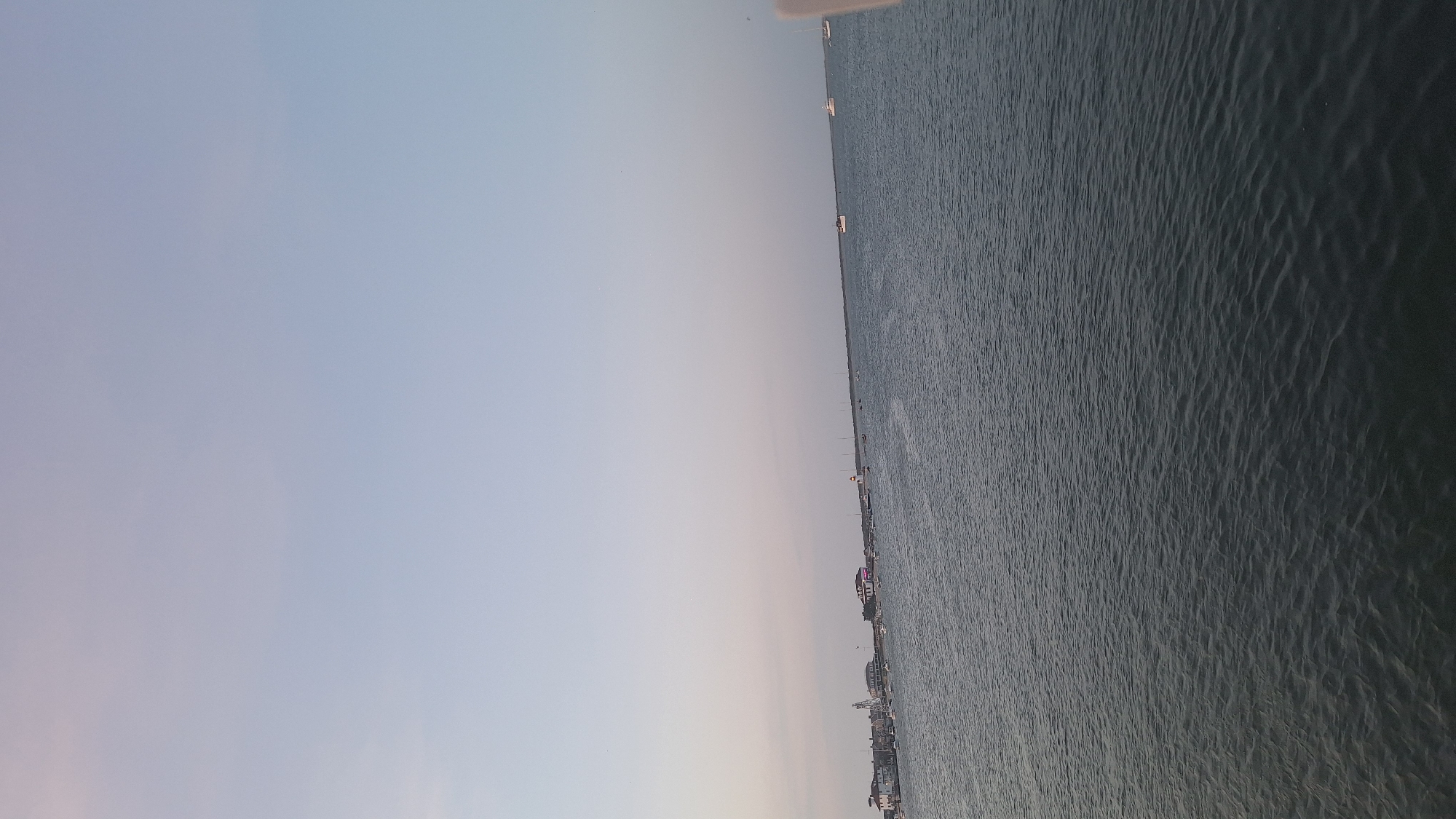 The image depicts a wide view of the ocean off the coast of Nantucket, Massachusetts, United States of America. The sky is a pale, cloudless blue, suggesting either early morning or late evening. The water is a deep, muted blue-green, with small ripples indicating a gentle breeze. In the distance, along the horizon, a line of buildings is visible, resembling a waterfront town or harbor. Several sailboats are moored or cruising on the water, varying in size and distance. A white lighthouse stands out among the buildings on the left side of the frame. The overall scene is tranquil and scenic, capturing the essence of a coastal environment.