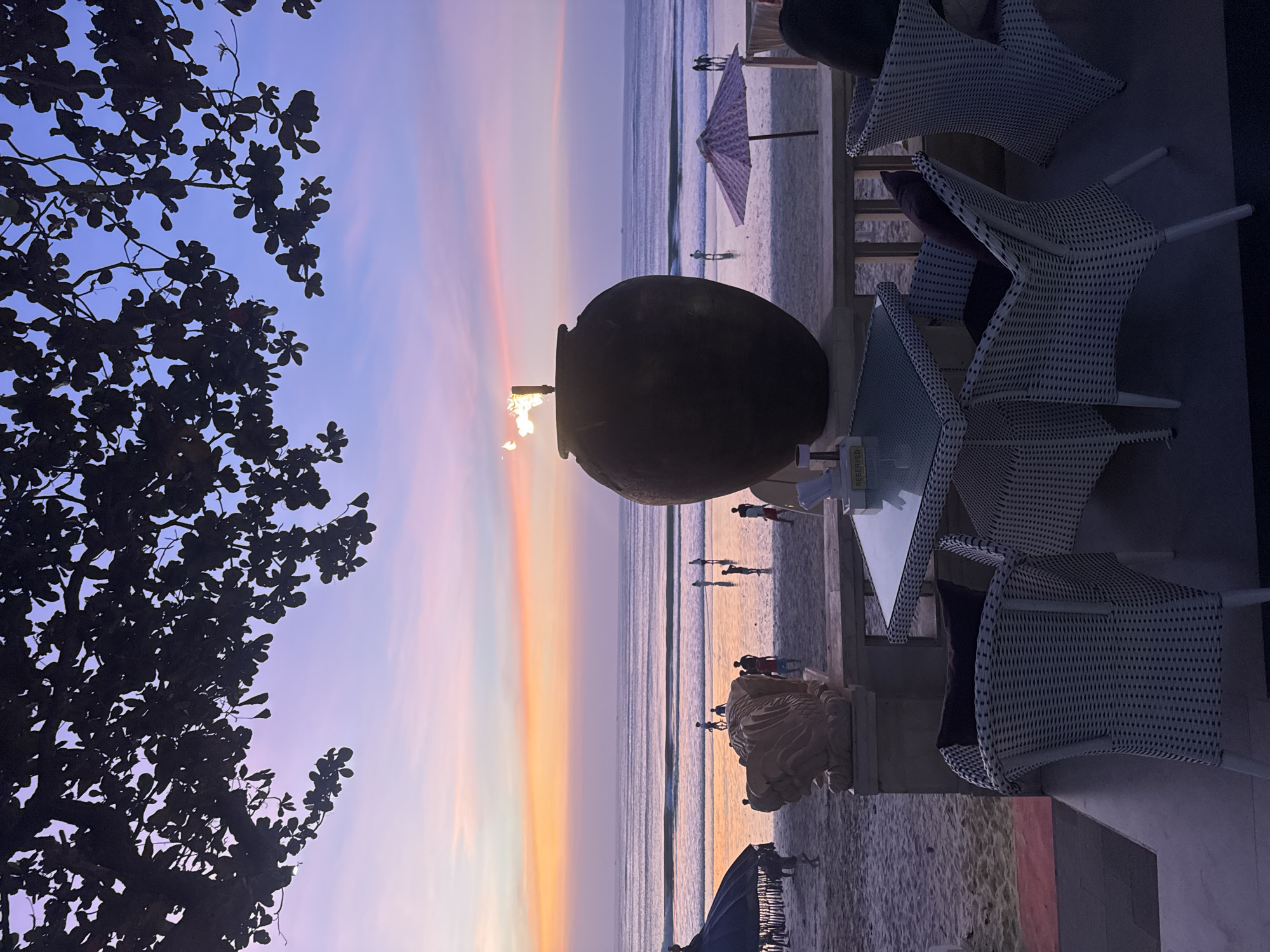 A wide-angle, eye-level, vertical shot shows a beachside dining area with the ocean and a sunset in the background. In the foreground, white wicker chairs and a glass-topped table are set up on a dark tiled patio. A large, dark, urn-shaped planter with a flame burning from the top sits on a ledge behind the table. The beach, with several people walking, is visible beyond the patio. In the distance, the ocean stretches to the horizon under a colorful sky with hues of purple, pink, and orange. The scene is likely Kuta Beach in Kuta Selatan, Bali, Indonesia, a popular tourist destination known for its sunsets and beachfront restaurants.