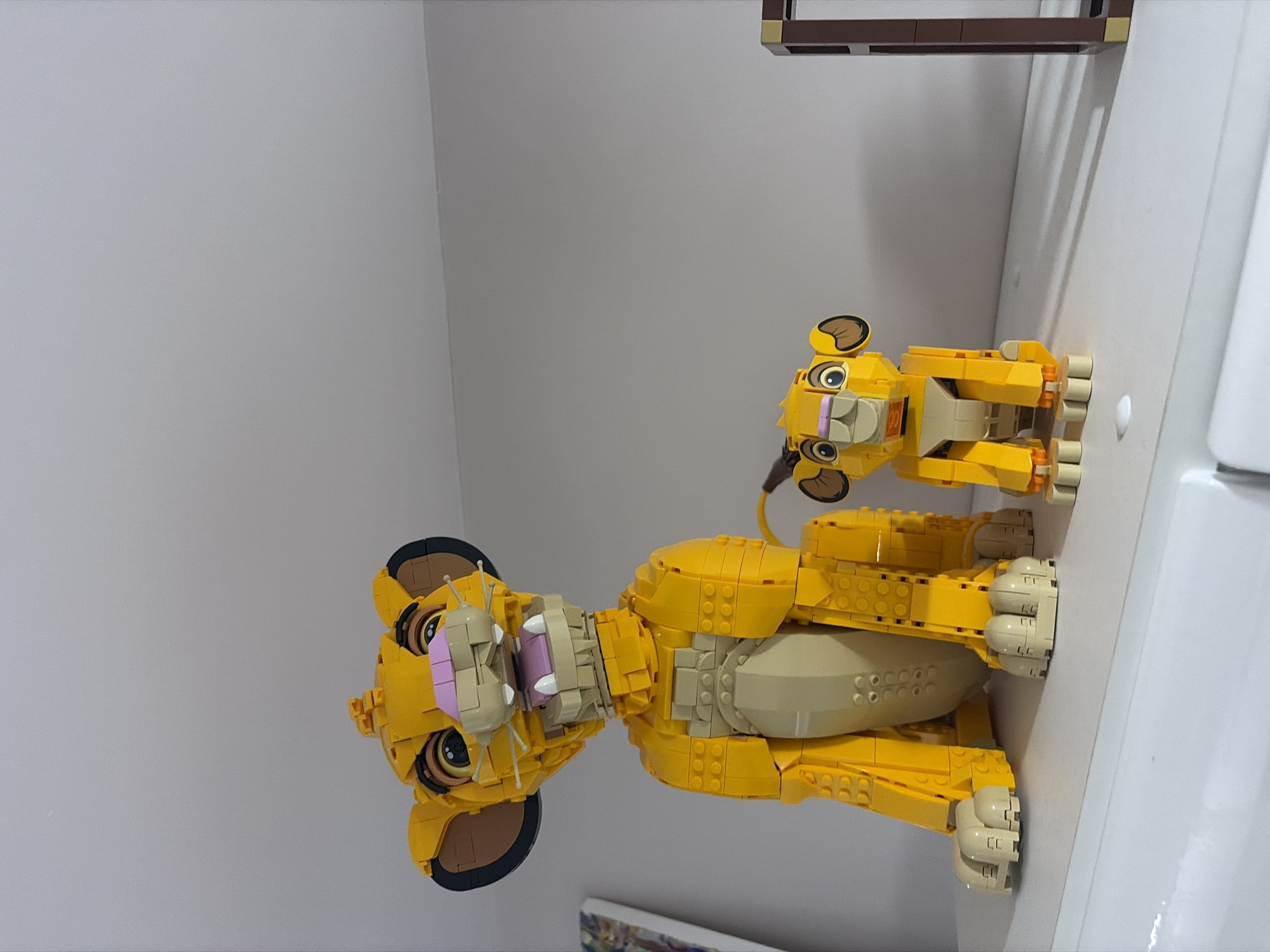 Two Lego lion figures, one significantly larger than the other, are displayed on a white shelf. The larger figure, positioned on the left, is a detailed replica of an adult lion, with yellow and tan bricks forming its body, head, and limbs. Its mouth is open, revealing pink and white interior details. The smaller figure, to the right, is a cub-like lion, also constructed from yellow and tan Lego bricks, sitting upright with its front paws resting on the shelf. The background consists of a plain, light-colored wall. The image does not provide any information about the city of Dobrich, Bulgaria, or any specific location within it.