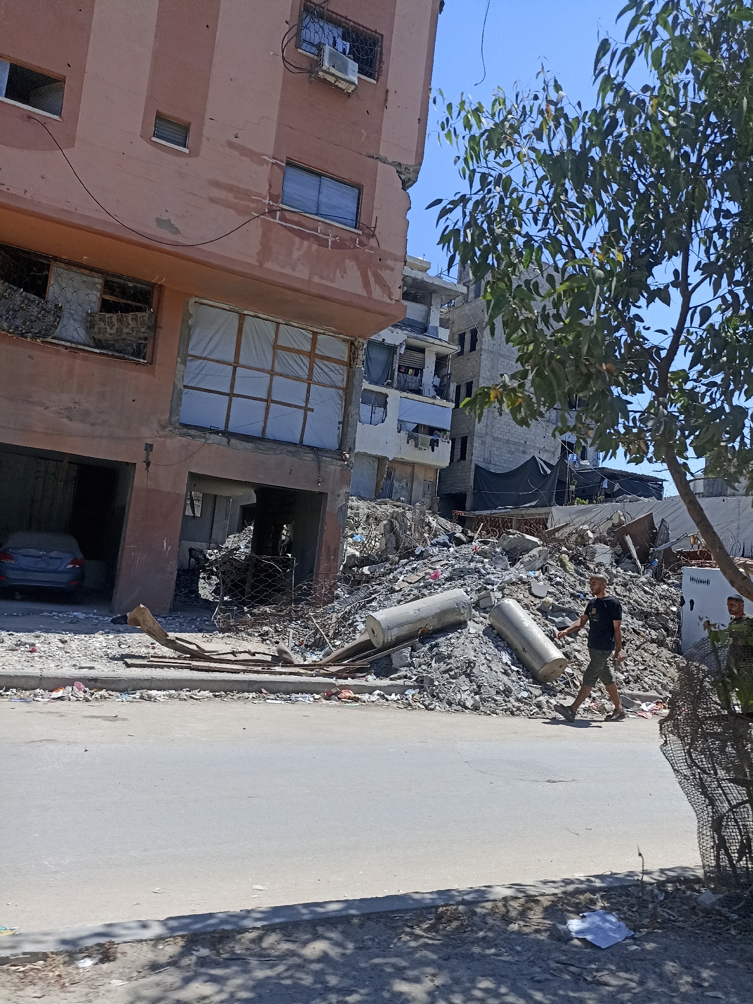 This is a medium shot of a street scene in Gaza City, Palestinian Territories, showing significant destruction. On the left, a partially damaged multi-story building, painted a muted pink, stands with exposed rebar and shattered windows. The ground floor appears to be a garage where a dark blue car is parked. To the right of this building is a large pile of rubble, composed of concrete, metal debris, and other construction materials. Two large, cylindrical concrete objects are visible within the rubble. A young man wearing a black t-shirt and olive green shorts is walking across the street towards the right side of the frame. Further to the right, another man, partially obscured by a wire fence, is visible. The street is paved and appears to be in a state of disrepair, with debris and trash scattered along the edges. In the background, other damaged buildings can be seen, some with tarps or coverings over windows. A tree with green leaves is positioned on the right side of the frame, casting shadows on the street. The sky is clear and blue.