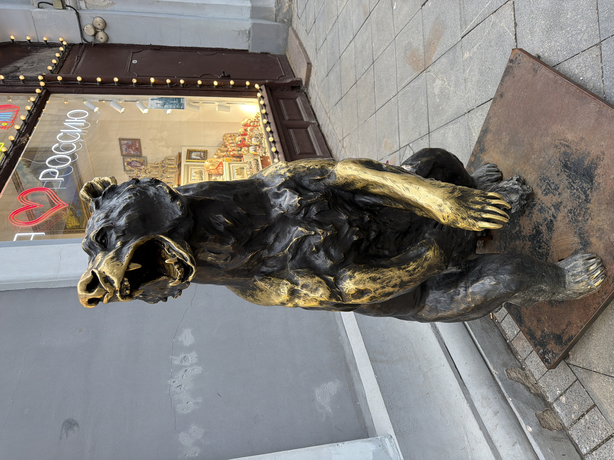 A large bronze statue of a bear dominates the foreground, depicted in a powerful, crouched pose with its mouth open, as if roaring or growling. The statue is primarily dark bronze, with significant portions of its fur, chest, front paws, and snout highlighted in a metallic gold finish, giving it a striking appearance. The bear rests on a rectangular, dark, weathered metal base.
In the background, behind and to the left of the bear, is a storefront with a large glass window. Inside the brightly lit shop, various objects, possibly souvenirs or decorative items, are visible on display. Above the shop window, a string of warm-toned light bulbs illuminates the facade. Visible through the window, in white neon letters, is the word "РОССИЮ" (ROSSIYU), which means "Russia" in Russian, accompanied by a red neon heart shape.
The building facade next to the shop is made of a light grey, textured material, possibly concrete or plaster. The ground is paved with grey, square-patterned tiles. The overall scene suggests an urban street environment during daytime, with the shop lights indicating it might be open for business. The image is oriented with the scene rotated 90 degrees clockwise, so the bear appears to be lying on its side.