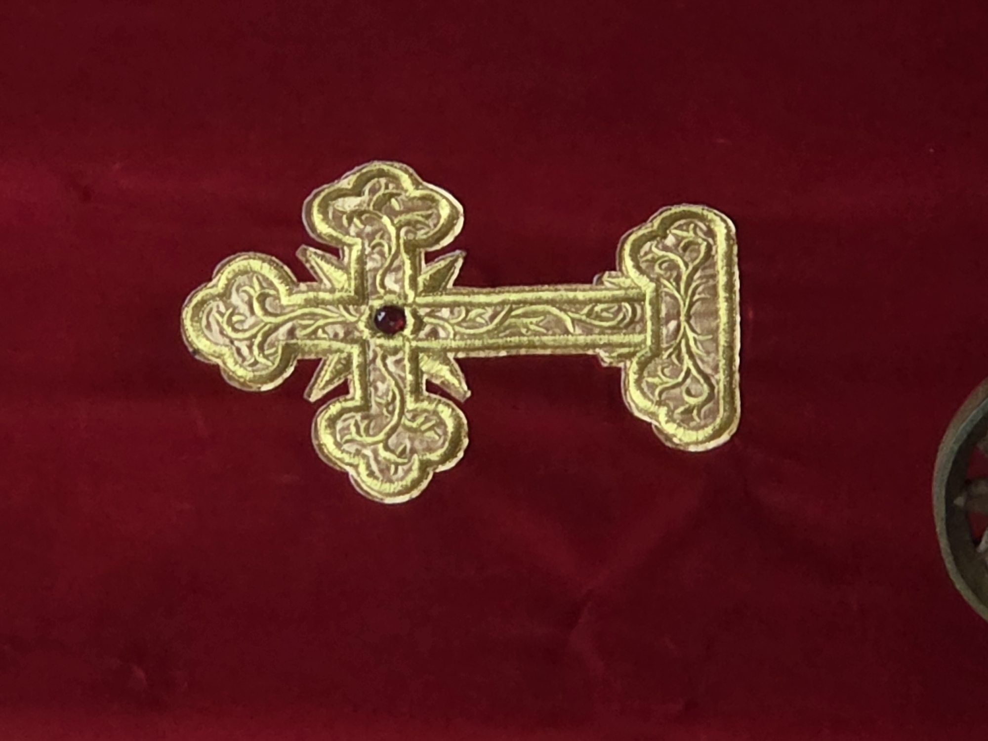 On a red surface, a gold Orthodox cross with a red gem at the center is positioned horizontally. Intricate scrollwork embellishes the cross. The location of Balsha, Bulgaria is not inferable from the image.