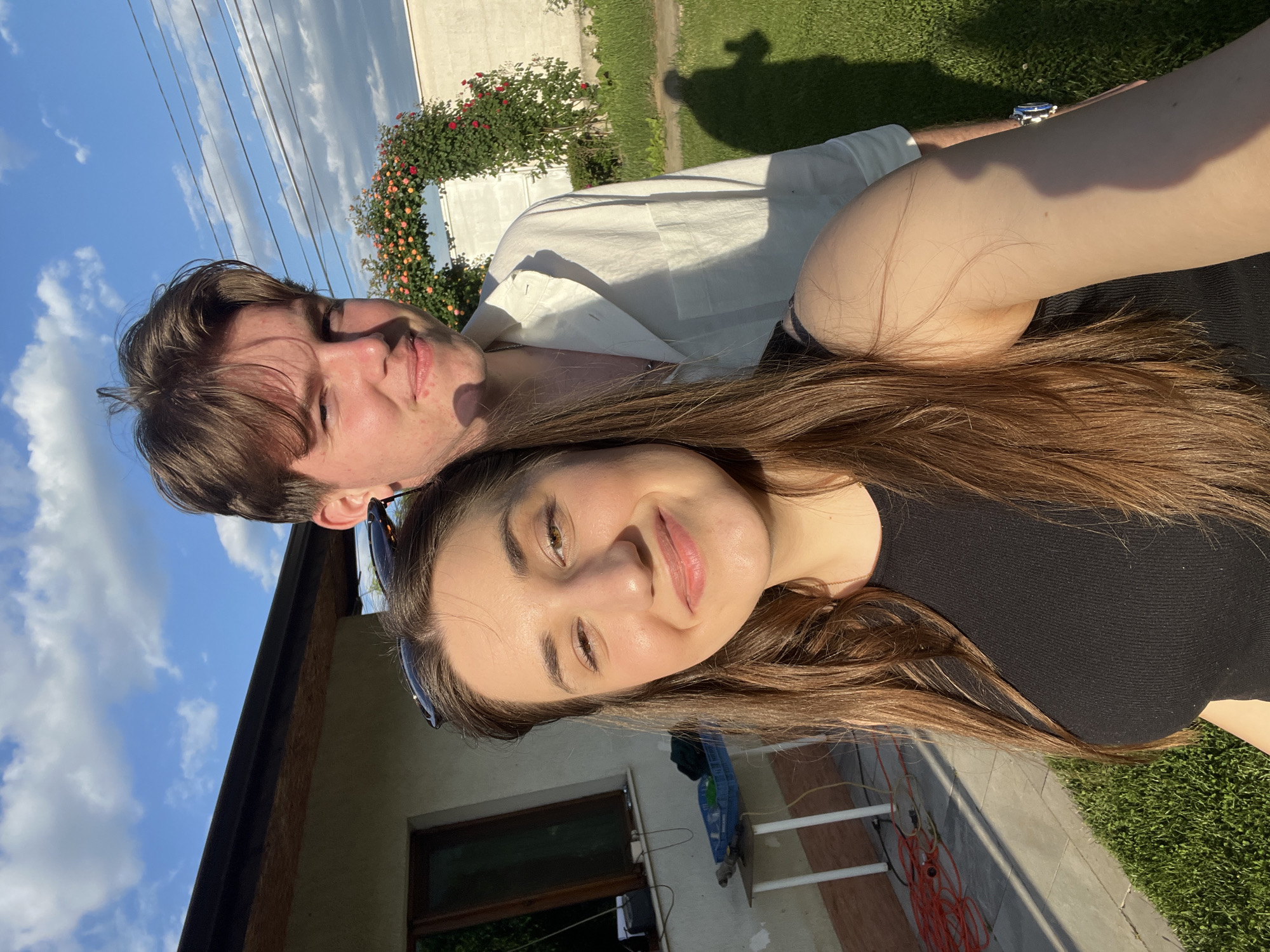 A young woman and man are posing for a selfie outdoors. The woman is in the foreground, wearing a black top, with long brown hair. Sunglasses are perched on her head. She is smiling. Behind her is a young man in a light-colored shirt. The background includes a building with a window, a rose bush, grass, and a blue sky with clouds. Power lines are visible in the upper left. The photo appears to have been taken in Busmantsi, Bulgaria. There is a shadow of a person on the grass.