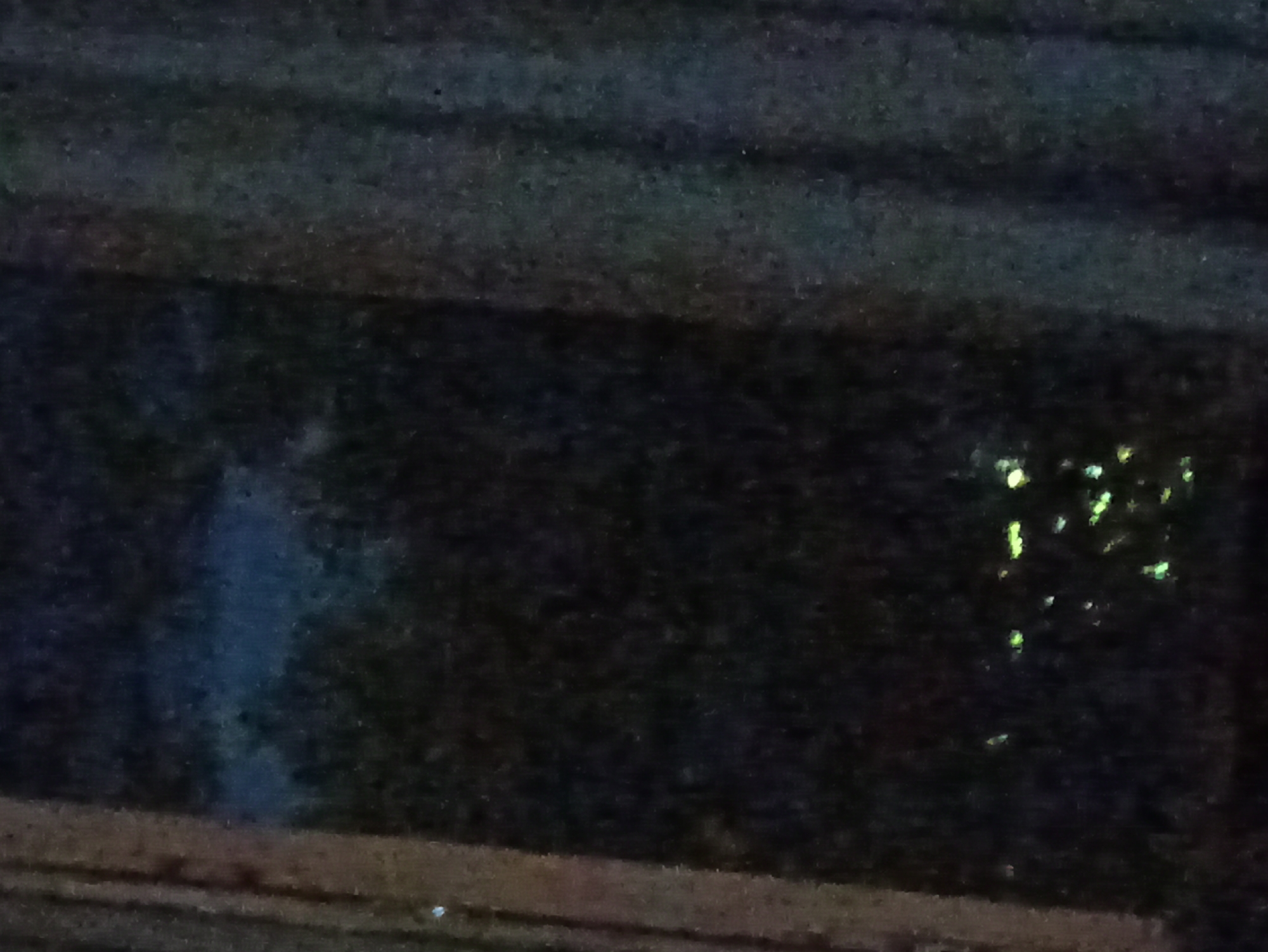 The image depicts a dark scene, likely taken at night. In the lower portion, there appears to be a ledge or baseboard. Above, a dark, undefined area is present, with a faint vertical blur on the left side and scattered, indistinct bright spots to the right, possibly reflecting light off of surfaces. The location is in a city of Unknown, Unknown.
