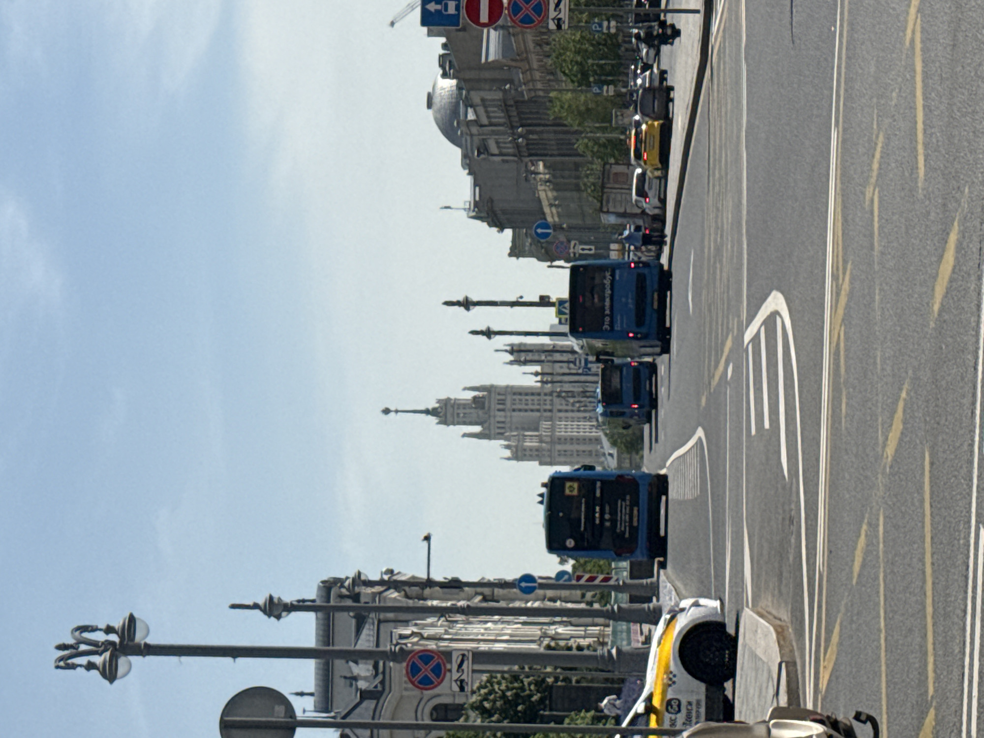 The image depicts a street scene in Moscow, Russia, featuring multi-lane road with traffic. Three blue buses are visible moving along the street, alongside a taxi with white and yellow livery. Pedestrian barriers line the side of the road, where street lamps are visible. In the background, a large building with a distinctive spire is recognizable. Traffic signs are present along the street. The sky is light blue, indicating daytime. The presence of specific architectural elements and traffic signs suggests that the image was likely taken along one of Moscow's main avenues.