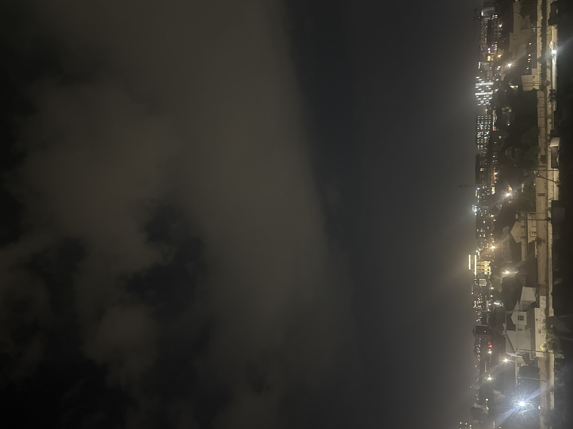 A night aerial shot captures a cityscape with a dark, cloudy sky overhead and illuminated buildings below. The clouds are dense and cover the upper portion of the frame, obscuring any stars or moon. The city lights are visible in the distance, creating a hazy glow that permeates the lower half of the image. Various buildings, including residential and commercial structures, are discernible, with some featuring illuminated windows. Streetlights also punctuate the darkness. The overall impression is of a sprawling urban area under a heavy, overcast sky at night. This appears to be a view of Bishkek City, Kyrgyzstan.