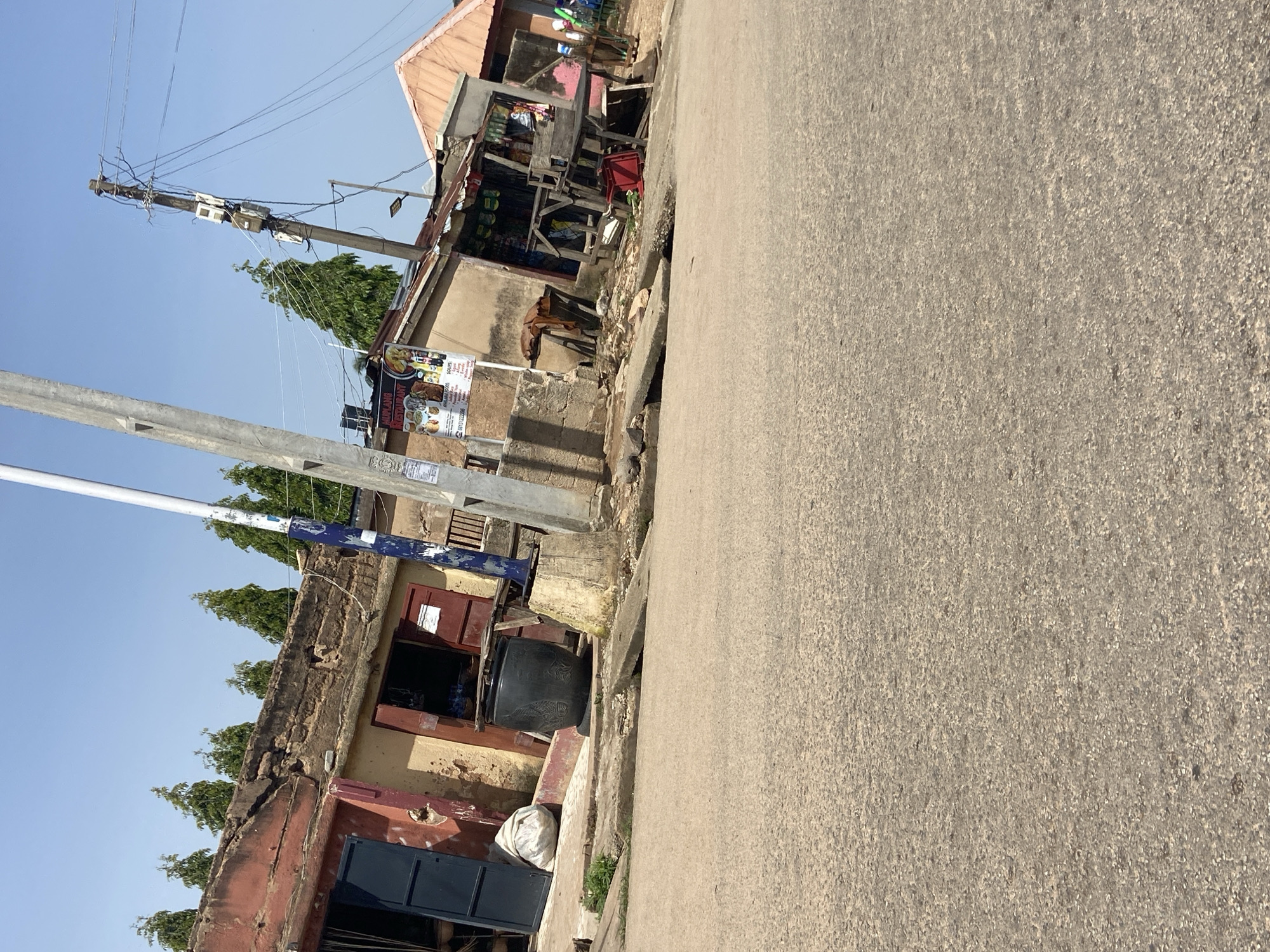 The image depicts a street scene in Jos, Nigeria, characterized by a row of aging commercial structures lining an asphalt road. The buildings exhibit signs of wear, with some walls appearing cracked and roofs showing disrepair. Several shops are open, displaying various goods. A large, dark, cylindrical container sits prominently in front of one of the shops, next to a sack of what appears to be white fabric or material. A utility pole with multiple electrical meters and wires stands tall, indicating the presence of electrical infrastructure. Trees with conical shapes are visible above the buildings, suggesting a moderately vegetated environment. The sky is clear and blue, indicating a bright, sunny day. The overall impression is one of a developing urban area with functional, albeit somewhat dilapidated, commercial activity.