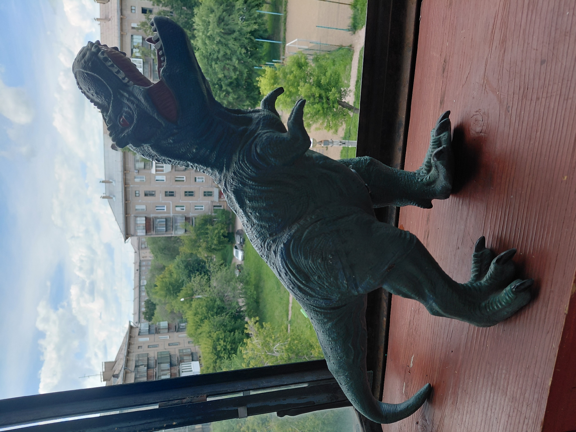 A dark green, toy Tyrannosaurus Rex figure stands on a reddish-brown wooden surface, positioned in front of a window overlooking a city. The dinosaur toy is in profile, facing right, with its mouth open and head tilted upward. The background visible through the window shows multi-story apartment buildings, lush green trees, and a cloudy sky. A sports field with a fence can be seen in the distance. The scene is likely located in Magnitogorsk, Russia, given the architectural style of the buildings. The lighting suggests it is daytime, possibly late morning or early afternoon.