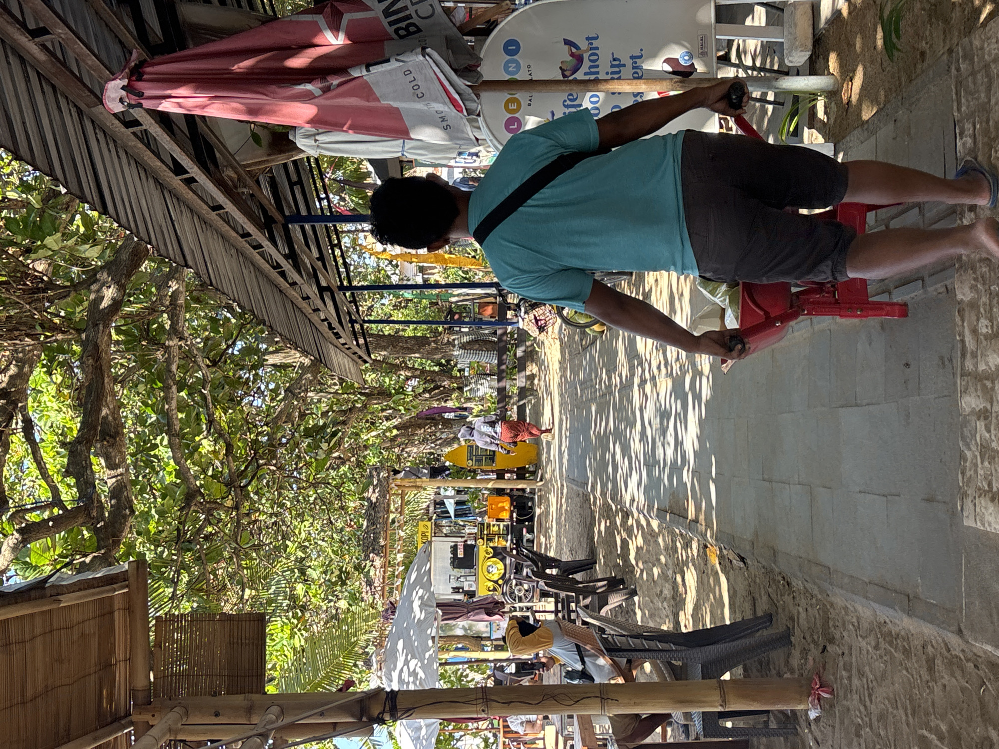 The image depicts a man pushing a red wheelbarrow on a tiled pathway at a beachside establishment in Denpasar, Indonesia. The man, seen from behind, wears a teal t-shirt and dark shorts, with a black strap diagonally across his chest. He is pushing the wheelbarrow, which contains what appear to be coconuts or similar items, and is barefoot with blue flip-flops.
The pathway is bordered by sand on the left, with several tables and chairs set up, some under a bamboo-covered structure. White umbrellas are visible, providing shade. Further back, a yellow food truck with "KOPI" signage is present, along with other structures and trees. On the right side of the pathway, a large red umbrella is partially furled, leaning against a structure. Behind the umbrella is a white circular sign with blue lettering that appears to be an advertisement. The environment is sunny, with dappled light filtering through the trees. The overall scene suggests a vibrant, casual atmosphere at a beach resort or cafe.