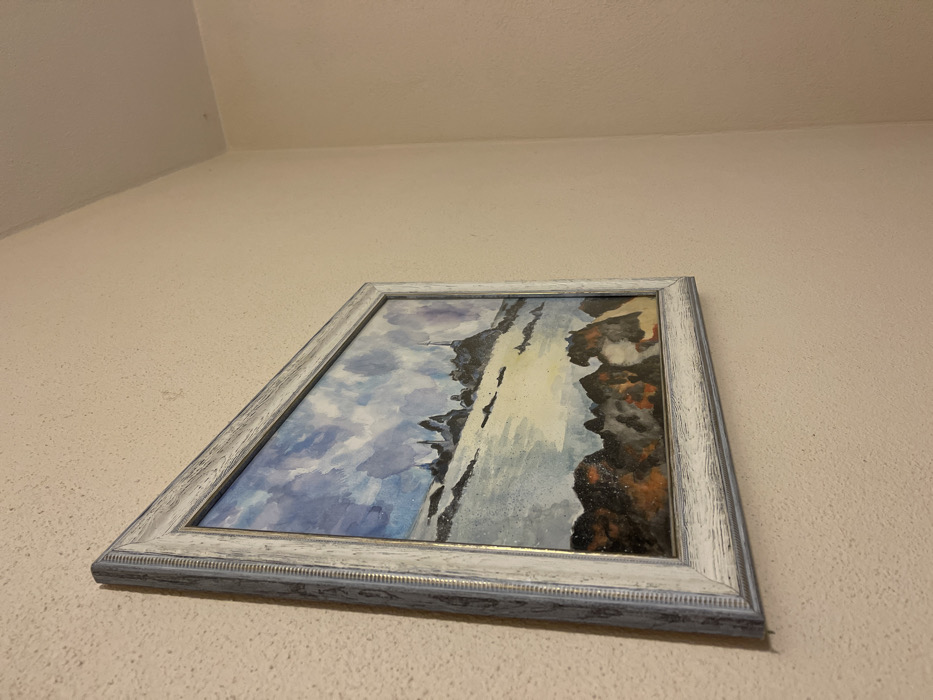The image displays a framed watercolor painting hanging on a textured, light-colored wall, captured from a low, upward-angled perspective.

The painting depicts a dynamic landscape, likely a dramatic waterfall or a steep coastal scene. The left side of the artwork features a cloudy sky rendered in blues and grays, transitioning into a body of water. The right side showcases dark, rugged land formations or cliffs in earthy tones of brown, orange, and black, possibly with sparkling or speckled accents. A prominent vertical streak of light colors, potentially a waterfall or reflections, bisects the composition.

The artwork is encased in an ornate, distressed wooden frame with a weathered white and light grey finish, accented with subtle blue-grey details and a thin gold-colored inner rim. The wall behind the painting has a granular or stucco-like texture and is a uniform light beige or off-white color. The corner where the wall meets the ceiling is visible in the upper portion of the photograph.

There are no people or specific activities visible in the image. The setting appears to be indoors, under ambient lighting, which does not indicate a specific time of day. No text is discernible anywhere in the frame or on the wall. While the location context is provided as Torre di Santa Maria, Italy, there are no specific visual elements within the photograph itself that confirm this location.