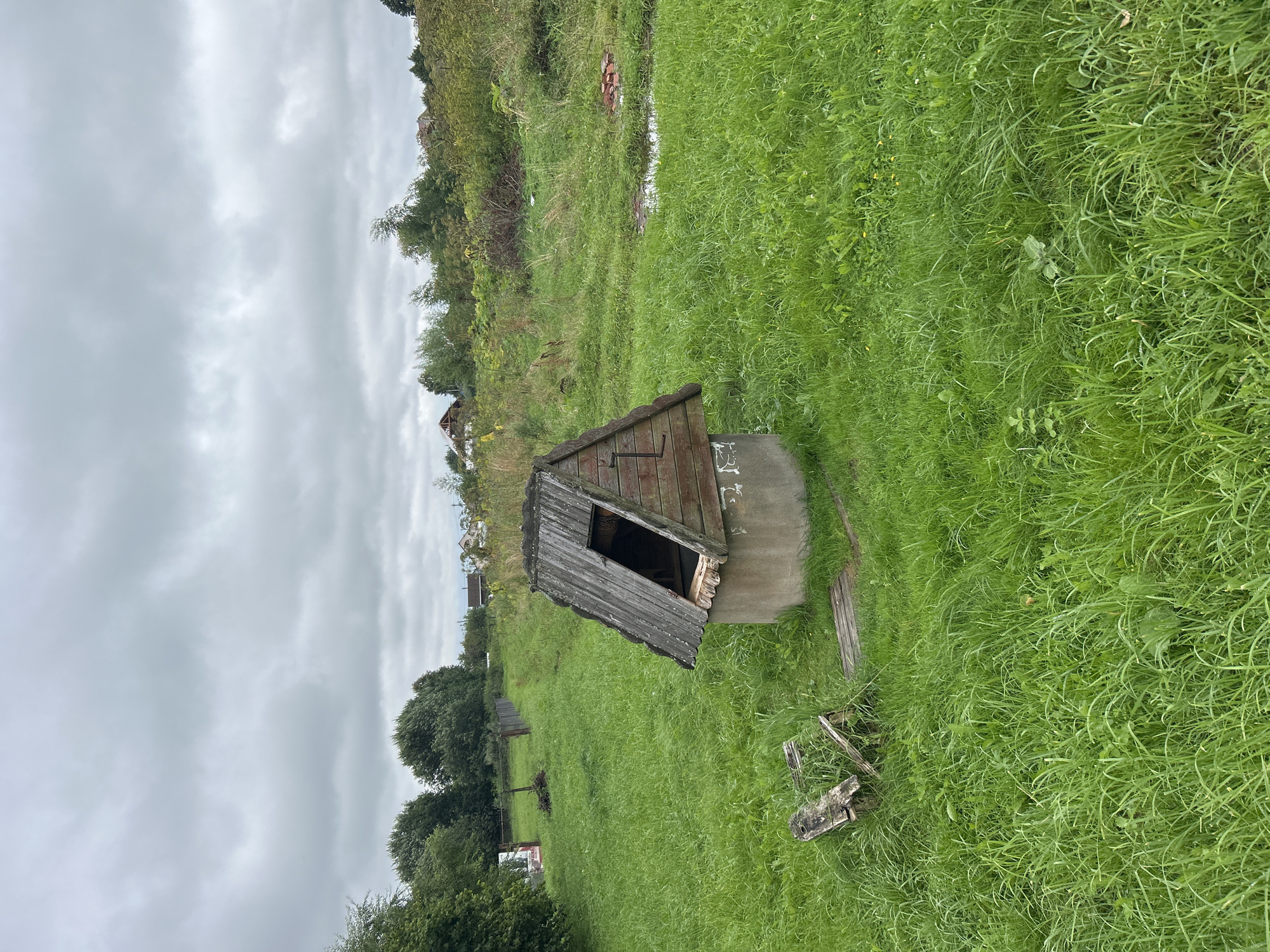 In the village of Kalitsino, Russia, an old, dilapidated wooden well with a broken roof stands in a grassy field. The well has a stone base and a weathered wooden structure on top, with one side of the roof missing. Next to the well, a broken wooden bench or seat lies on the ground. The surrounding area is covered in tall, green grass, and in the distance, trees and some houses are visible under a cloudy sky. The scene appears to be in a rural, overgrown setting.