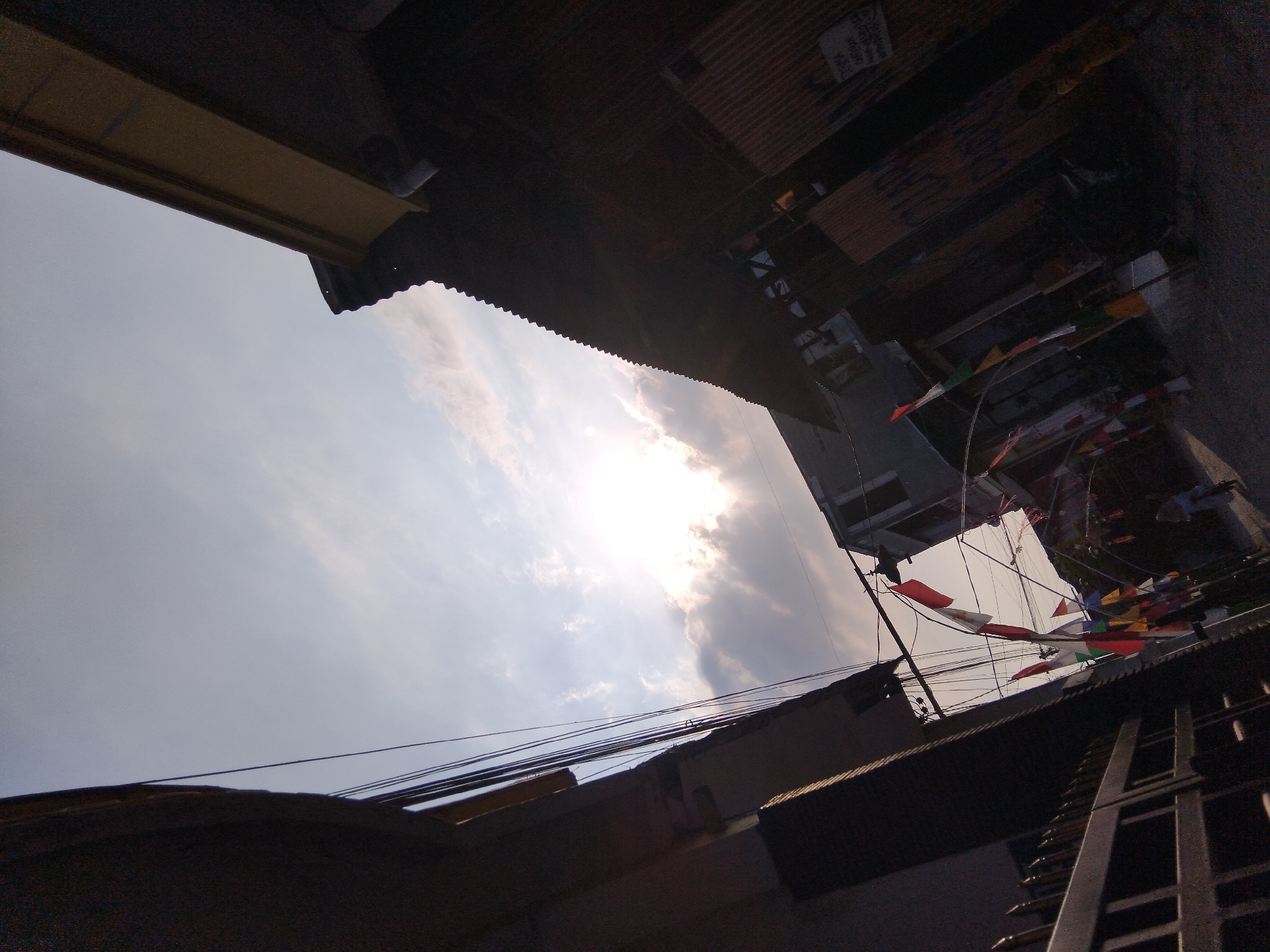 The image is a low-angle shot of a street in Kepuh Kiriman, Indonesia, with buildings on either side and a view of the sky. The sun is shining brightly through clouds, creating a lens flare effect. Various flags, including what appear to be Indonesian flags, are strung across the street, suggesting a festive or commemorative occasion. Wires are visible crisscrossing the sky above the street. The buildings are of varying heights and materials, with one on the left appearing to be a residential building with a dark facade, and another on the right constructed with corrugated metal roofing and siding. A metal fence is present on the left side of the image. In the distance, a few figures are visible walking or standing on the street. The scene suggests a typical urban or suburban environment in Indonesia, possibly during a celebration.