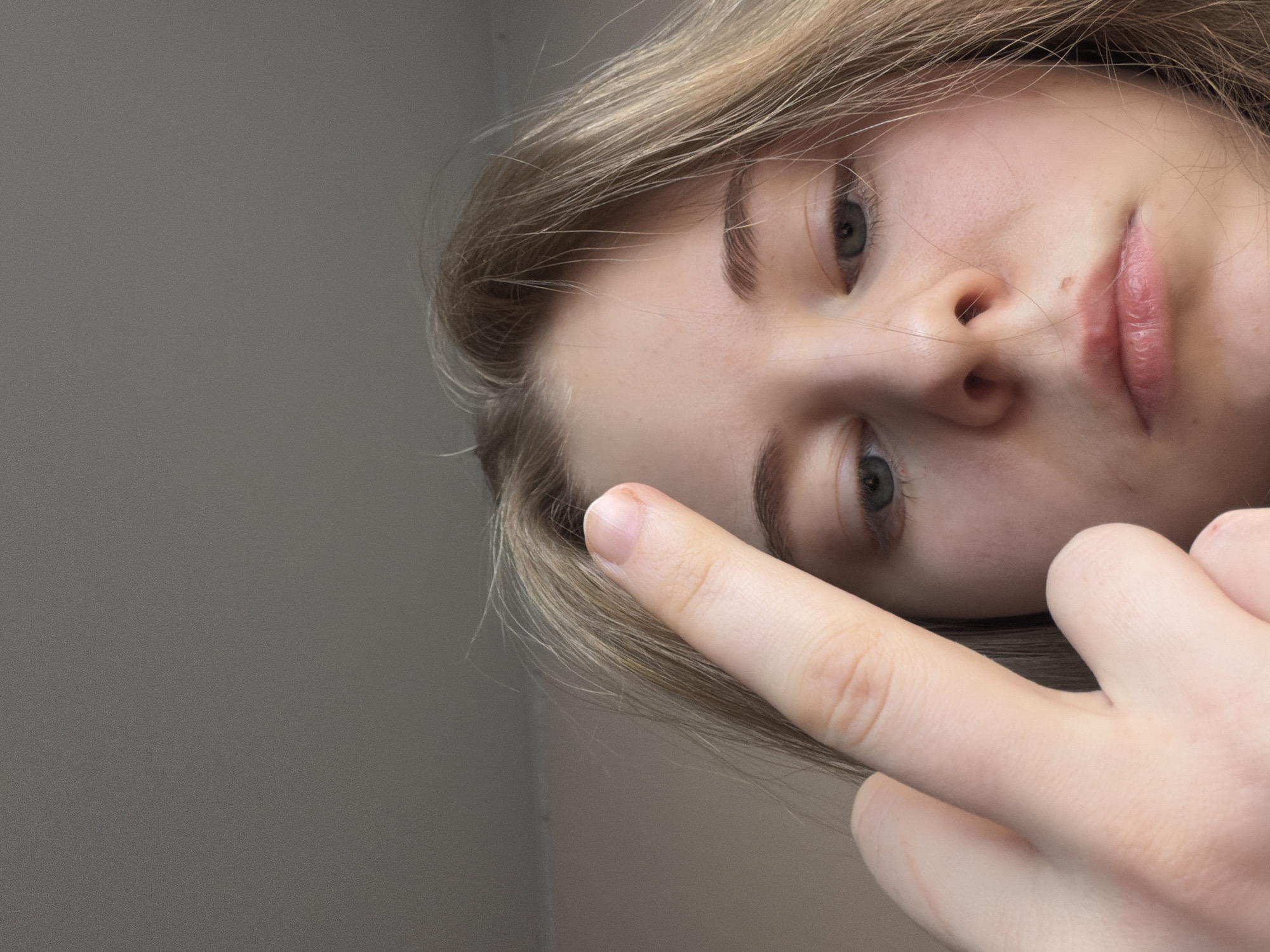 The image is a close-up of a person's face, angled as if looking down at the viewer. The person has fair skin, light brown hair, and gray eyes. The person's middle finger is extended towards the viewer, partially obscuring the face. The background is a plain gray wall. The location in the city of Perm, Russia is not discernible from the image. No city-specific landmarks are visible.