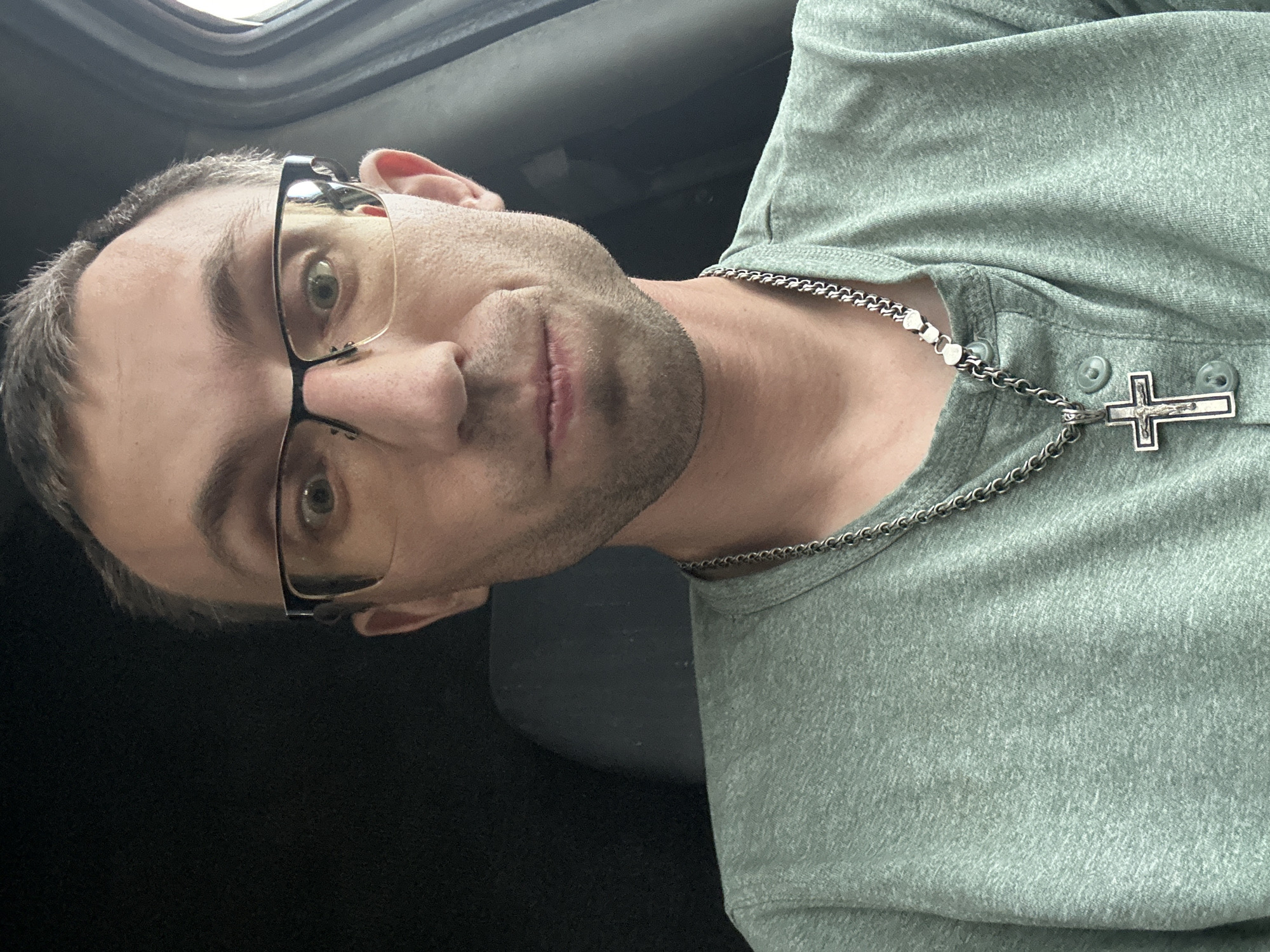 A man in his 30s, wearing a pale green henley shirt and glasses, is photographed from the chest up. He is looking directly at the camera with a neutral expression. A silver chain with a cross pendant rests on his shirt. The background is dark, suggesting an interior space, possibly a vehicle, with a glimpse of a grey seat on the left. There is no discernible location within the city of Berezivka, Ukraine.