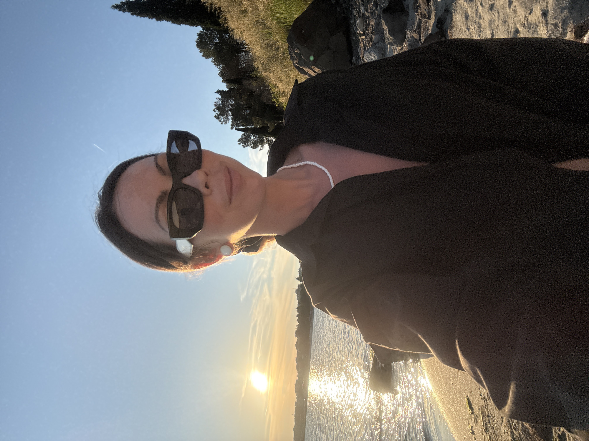 A medium shot selfie shows a woman on a rocky beach in Nesebar, Bulgaria, at sunset. The woman is wearing large black sunglasses, a dark unbuttoned shirt, and a white beaded necklace. Her hair is pulled back. The sun is setting over the water, casting a warm glow on the scene. The background features trees and vegetation on a hillside. The foreground is a sandy beach with rocks.