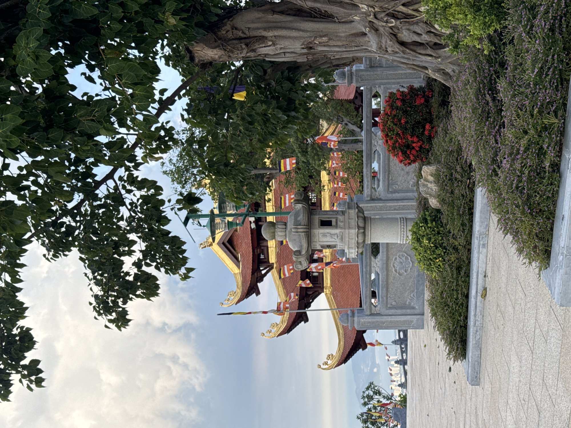 The image depicts a multi-tiered Buddhist pagoda with red tiled roofs and golden accents, situated in the city of Diên Khánh, Vietnam. The pagoda is partially obscured by lush green foliage from a large tree with exposed roots in the foreground, and other trees and bushes. Prayer flags in various colors are visible, adorning the pagoda and surrounding structures. A stone lantern stands prominently in front of a decorative stone wall. The ground is paved with light-colored square tiles. The sky is partly cloudy.
