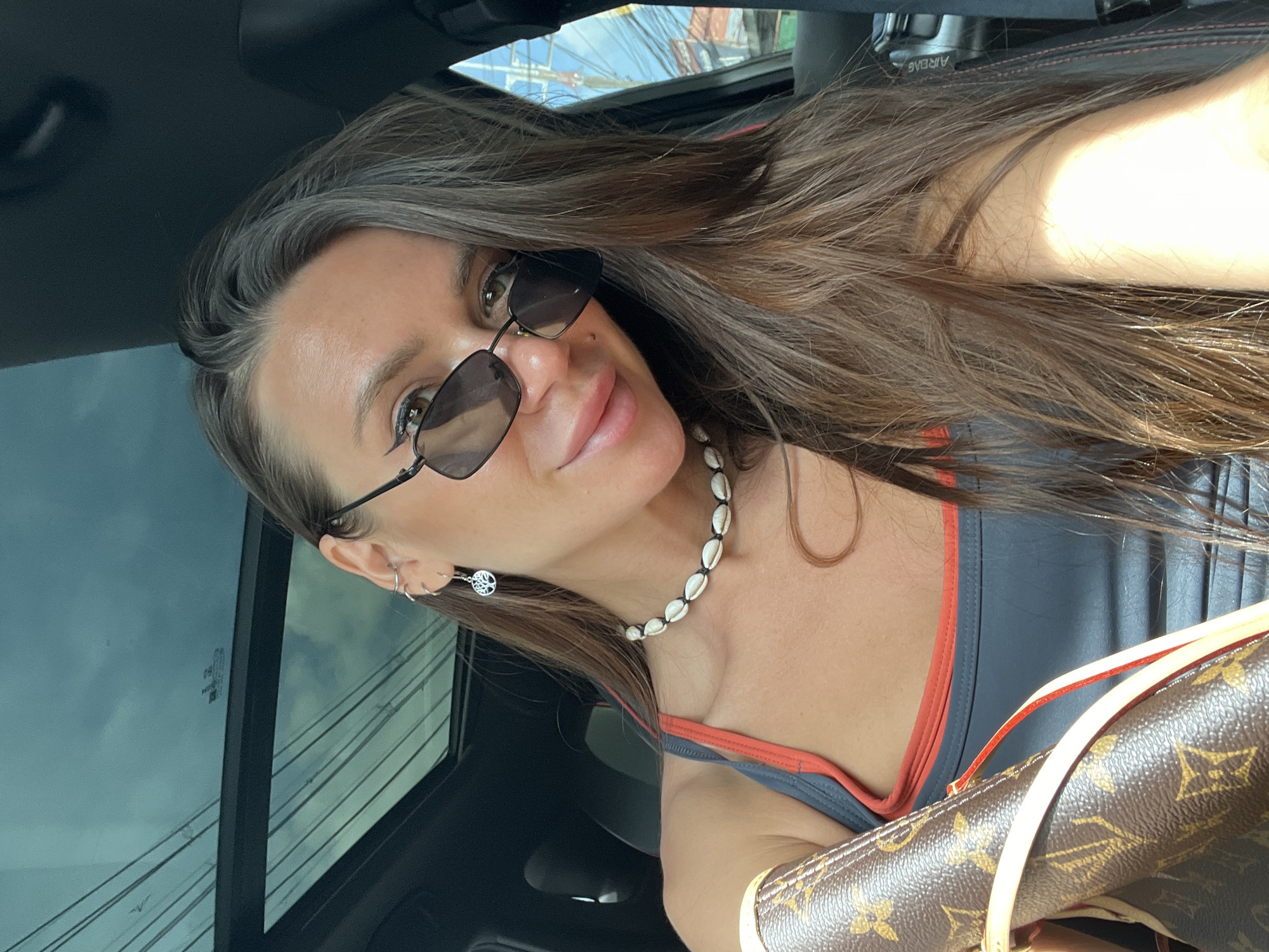 A woman with long, brown hair, wearing sunglasses, a shell necklace, and a tank top, sits in the driver's seat of a car with a sunroof open. A designer handbag is visible on her left side. The background shows buildings and power lines, suggesting an urban or suburban environment. The interior of the car is dark. The lighting indicates it is daytime. The specific location of Baan Bo Phut, Thailand is not discernable from the visual information in the image.