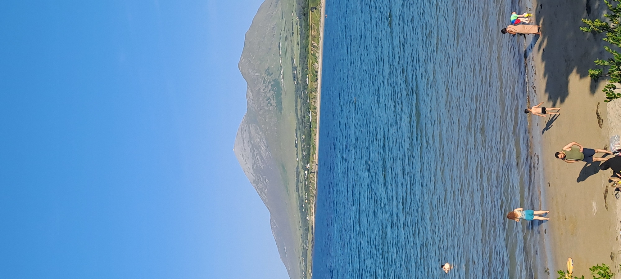 The image depicts a coastal scene with a large mountain in the background. The sky is a clear, bright blue, and the water is a deep blue with gentle waves. In the foreground, there is a sandy beach with several people. A man in a green tank top and blue shorts stands on the sand, facing to the left with his hands on his hips. A child in black swim trunks walks away from the water. Further to the right, two adults, one in a light-colored dress and the other in colorful clothing, appear to be interacting with a child in a blue swimsuit. Another child with red hair and blue shorts stands at the water's edge. In the water, a person is partially submerged. The mountain in the background is large and appears to be covered in a light-colored, rocky surface, with green hills and scattered houses at its base. The overall impression is a sunny day at a beach with a prominent natural landmark.