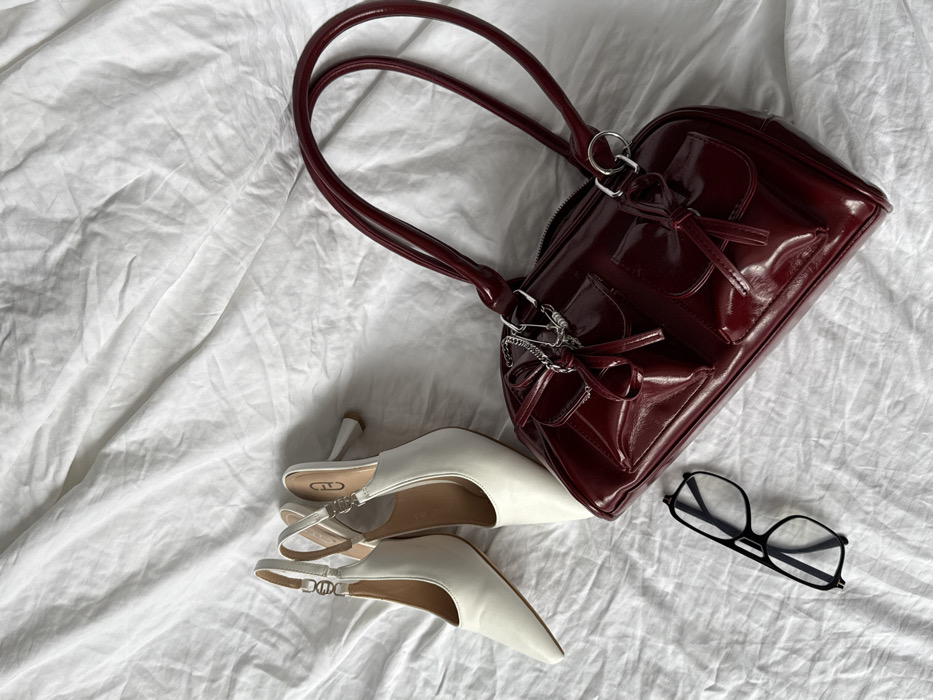 The scene depicts a static arrangement of personal accessories on a white, wrinkled fabric surface, consistent with a rumpled bedsheet, located in the city of Krasnodar, Russia. In the upper-left quadrant, a pair of white, pointed-toe slingback heels with mid-height heels are positioned, one slightly overlapping the other. Each shoe features a thin heel strap adorned with a small, silver-toned chain-link embellishment. To the right and slightly below the shoes, a burgundy, patent-leather finish shoulder bag is placed diagonally across the mid-right portion of the frame. This bag has two top handles, a visible top zipper, and two structured flap pockets on its front, each secured with a small, decorative bow-shaped tie made from the same material. A metallic silver-toned chain with several interlocking rings is attached to one of the bag's handles. In the lower-left foreground, a pair of black-framed eyeglasses with clear lenses rests on the fabric. No individuals are present in the image.