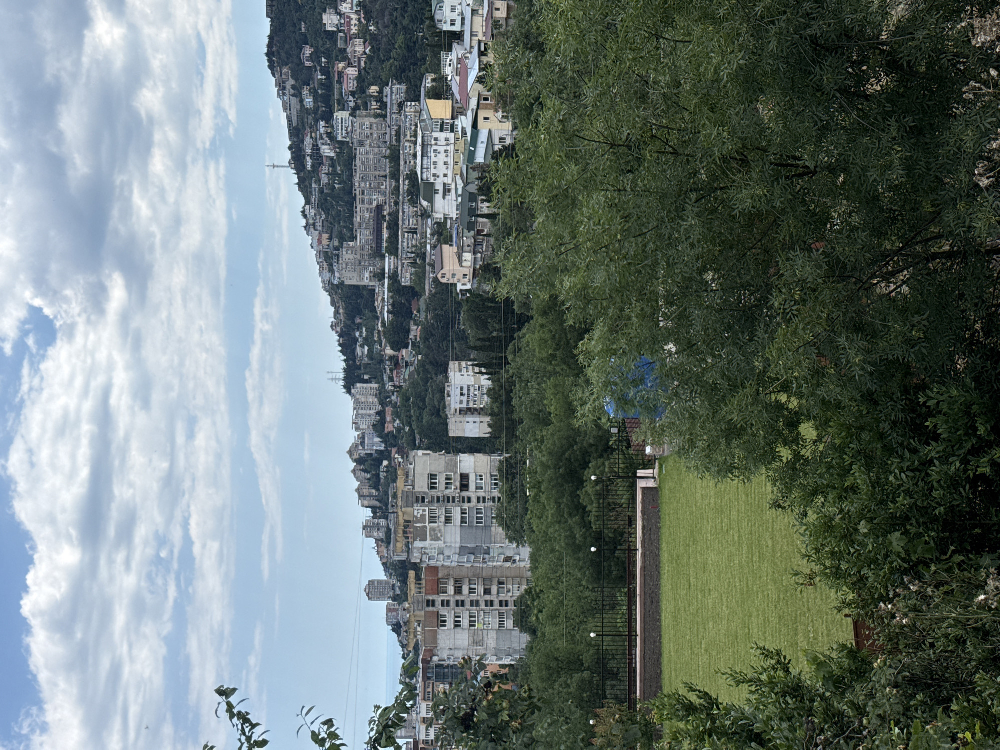 This is a vertically oriented, medium-shot, eye-level, outdoor photograph taken on a bright, cloudy day. The scene appears to be a coastal city built on a hillside, likely Yalta, Ukraine, given the context. In the background, a dense cluster of multi-story buildings, varying in architectural style and color, ascends the steep incline of a mountain covered in lush greenery. Some buildings have red-tiled roofs, while others are painted white or light gray. A tall transmission tower is visible in the distance, cresting the hillside.
In the foreground and mid-ground, the image is dominated by dense foliage from trees, with their leaves casting shadows on the ground. A section of a manicured, bright green lawn occupies the lower right quadrant of the frame, bordered by a black metal fence. The lawn appears to be artificial turf. Several white globe lights are strung along the fence line. The overall atmosphere is serene, with no people or specific activities clearly depicted, suggesting a quiet day in a residential or recreational area overlooking the city. The time of day is likely midday, based on the bright, diffused light characteristic of a cloudy sky. There is no visible text in the image.