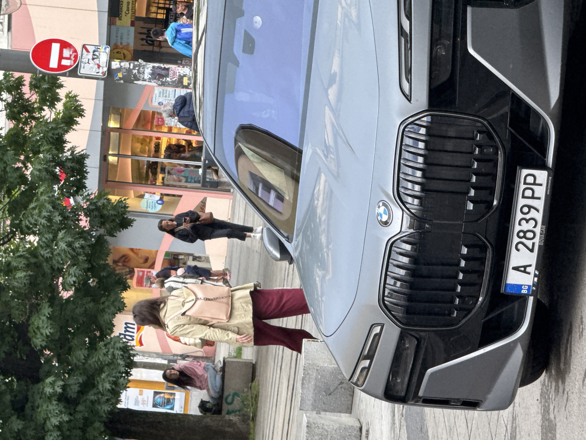 This image captures a vibrant daytime street scene in Sofia, Bulgaria, focusing on a modern silver/grey BMW car parked on the right side of a sidewalk. The car, possibly an SUV, features a distinctive large black grille and displays a Bulgarian license plate "A 2839 PP" which also includes "BG", "BOYA CAR", and "www.bovacar.bg". Reflections on the car's windshield show architectural details of surrounding buildings.
Several people are visible engaged in casual activity. In the mid-ground, a person wearing a light beige jacket and maroon pants, carrying a light pink shoulder bag, walks on the sidewalk. Further back, another individual in a dark jacket stands, appearing to look at a phone. A person in a blue jacket is partially obscured behind the car. To the far left, a person in a pink top is seated on a low wall or bench.
The background reveals a bustling urban environment with commercial buildings. Visible storefronts and signs include "dm" (likely a retail chain), "Испанска" ("Spanish"), "Санчо" ("Sancho"), and "RESTAURANT," suggesting a diverse commercial area. Another sign with "99%" is also visible, along with the word "ЖЕН" (female/women), possibly part of an advertisement. On the left, a prominent red circular "No Entry" sign is affixed to a pole, amidst the green foliage of a tree. The weather appears bright, possibly overcast, typical of a city day. The scene depicts everyday life on a busy street.