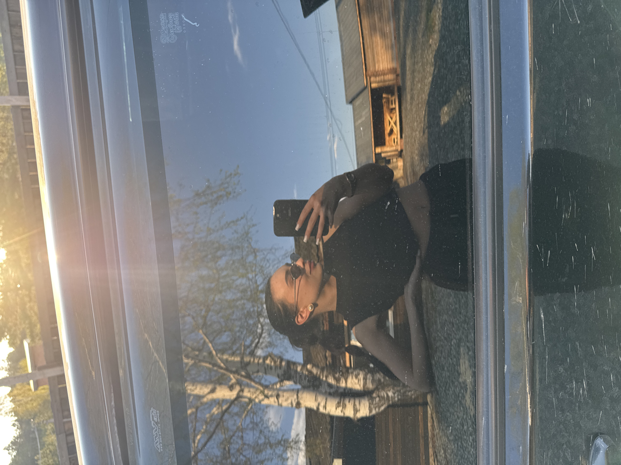 The image shows a reflection in a car window. The reflection includes a woman holding a smartphone, wearing sunglasses, a black top and bottoms, and jewelry. The reflection also shows bare trees, a clear blue sky, and a building with wooden siding and visible framework. The lighting suggests it is near sunrise or sunset. The location is specified as the city of Chaikovsky, Russia. The visible scratches and streaks on the car window obscure some details of the reflection.