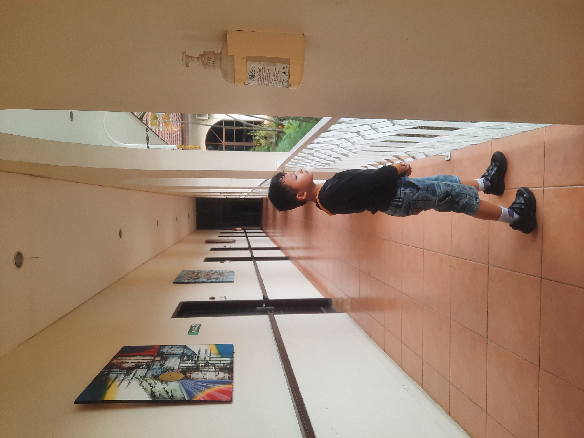 A young boy stands in a covered outdoor hallway, his back to the viewer, looking out past a white railing at a green, leafy area. He is wearing a black long-sleeved shirt, denim shorts, white socks, and black shoes. The hallway floor is tiled in a terracotta color. To the left of the boy, the hallway continues, lined with white walls featuring framed artwork. Several doors are visible along this wall. On the wall to the right of the boy, a dispenser for hand sanitizer is mounted. The hallway opens up to a view of buildings with red tile roofs and trees. The scene appears to be a hotel or resort in Babakan Madang, Indonesia, given the tropical foliage and architectural style.