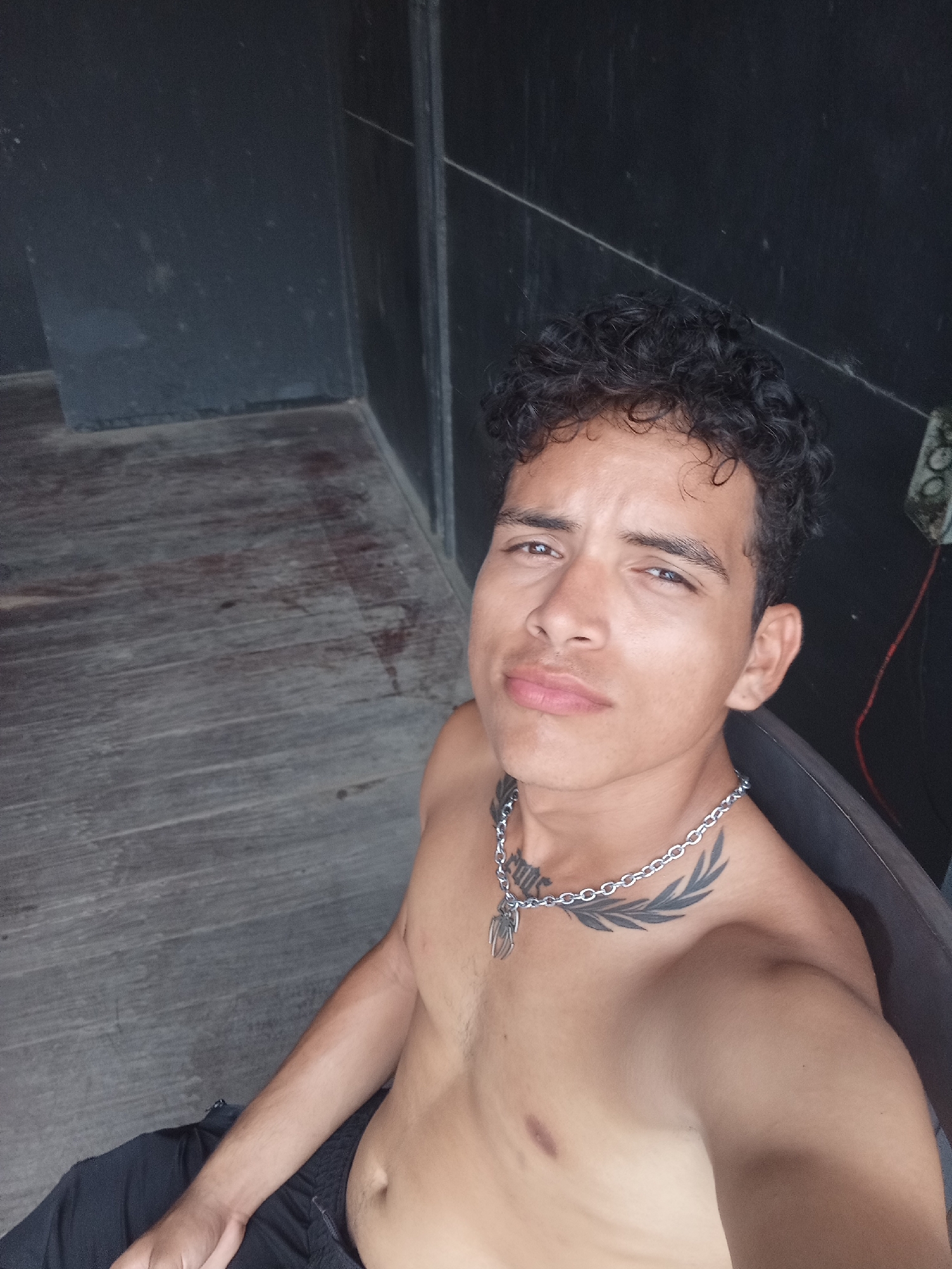 A shirtless young man with dark curly hair, a silver chain necklace, and a chest tattoo sits in a chair indoors. He is looking directly at the camera. The background consists of dark wooden flooring and dark walls with a vertical seam. A power outlet with wires is visible in the upper right corner. The location is assumed to be in Playa El Agua, Venezuela, based on the request.