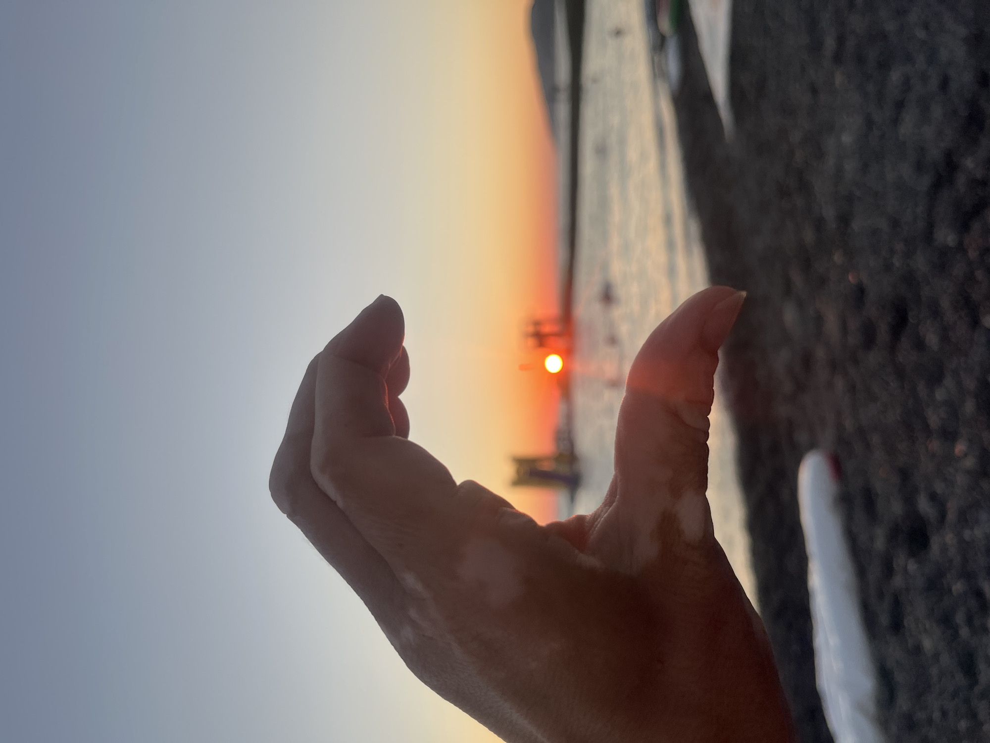A hand forms a heart shape with the setting sun in the background at a beach. The scene appears to be a coastal area with a body of water, a shoreline with dark pebbles, and a distant landmass. There are some blurred figures and structures in the water and on the beach. The sky is a gradient of pale blue and orange hues, indicating sunset. The hand in the foreground shows patches of vitiligo. The image could have been taken in Sochi, Russia, given the commonality of beaches and sunsets there.