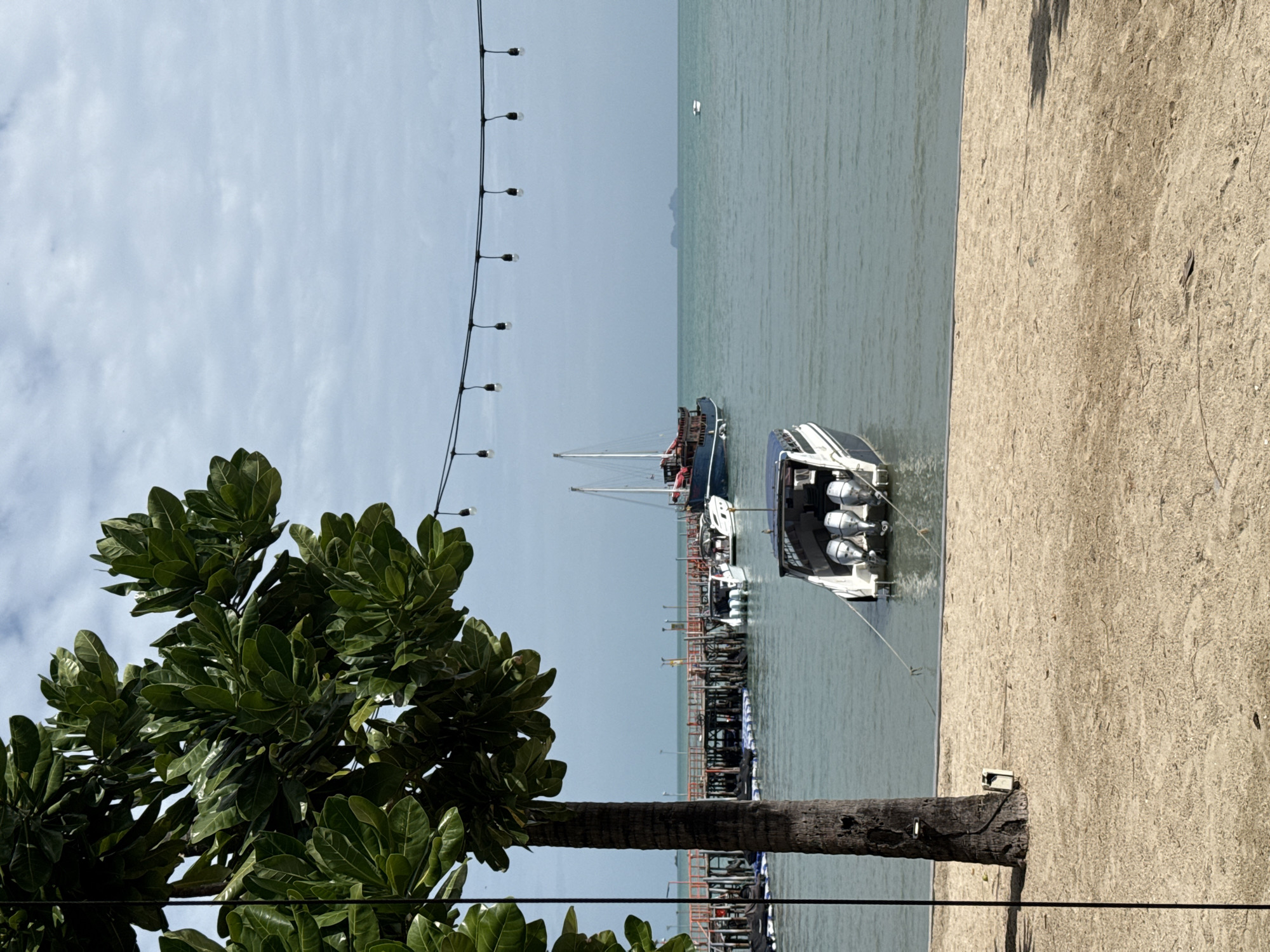 In Ko Samui District, Thailand, a beach scene unfolds with a sandy foreground and a calm, blue-green ocean. A palm tree stands on the left, its fronds partially obscuring the view of the sky, which is a muted grey with scattered clouds. A string of lights is suspended horizontally across the upper portion of the image, with visible bulbs.
In the water, several boats are present. A speed boat with three outboard motors is anchored in the mid-ground, close to the shore. Further out, a larger blue and red vessel, possibly a traditional Thai longtail boat or a similar fishing boat, is docked or anchored near a wooden pier. The pier extends into the water, with orange and brown wooden structures. More smaller boats and floating platforms are visible near the pier. In the distance, a sailboat with a white sail can be seen. The horizon is marked by a faint, hazy outline of a distant island. The overall atmosphere is tranquil, suggesting a serene coastal environment.