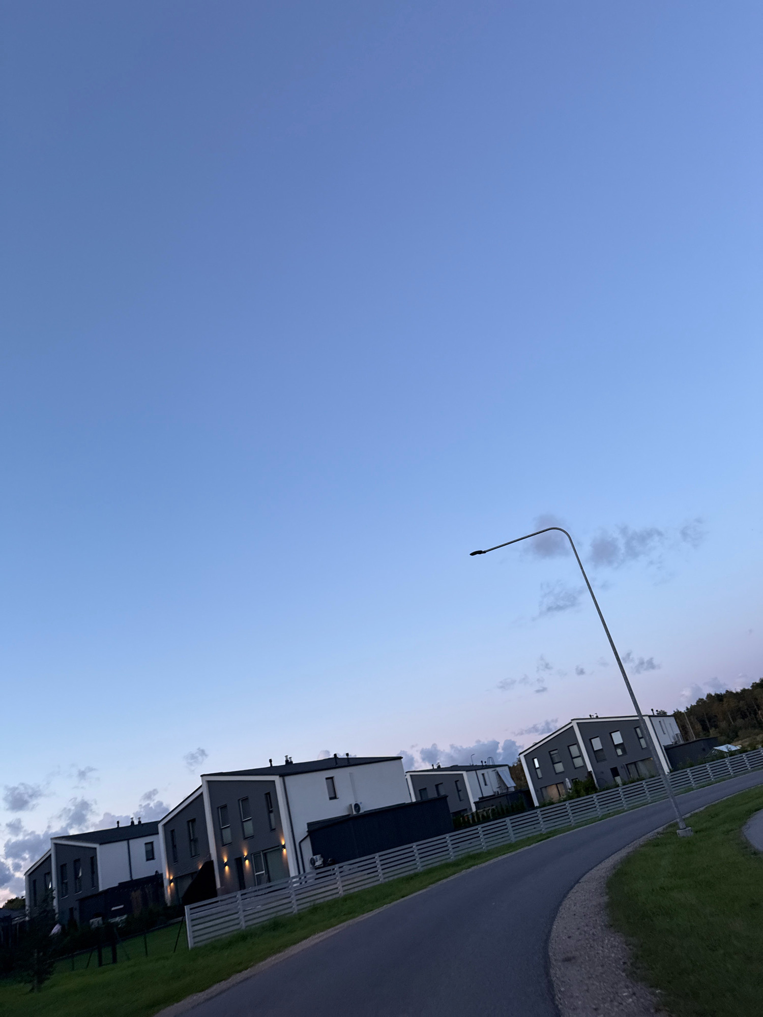 The image captures a suburban street scene in Alliku, Estonia, during twilight. The sky is a gradient of pale blue, transitioning to lighter hues near the horizon, with scattered, wispy clouds. A tall, modern street lamp with a curved arm is prominently featured on the right side of the frame, extending into the sky.
In the middle ground, a row of contemporary townhouses lines the street. These residences exhibit a modern architectural style with sharp angles, flat roofs, and a color palette of white, dark grey, and black. Many of the houses have large windows and exterior lighting that is illuminated, suggesting it is early evening. A light-colored, horizontal-slatted fence separates the properties from the street.
The street itself is paved and curves gently to the right. It is bordered by a well-maintained grassy area on the right, which includes a sidewalk or pedestrian path. On the left side of the street, more of the townhouses are visible, along with manicured lawns and some evergreen trees. In the background, a line of dark trees marks the edge of the development. The overall impression is one of a quiet, modern residential neighborhood. No people are visible in the image.